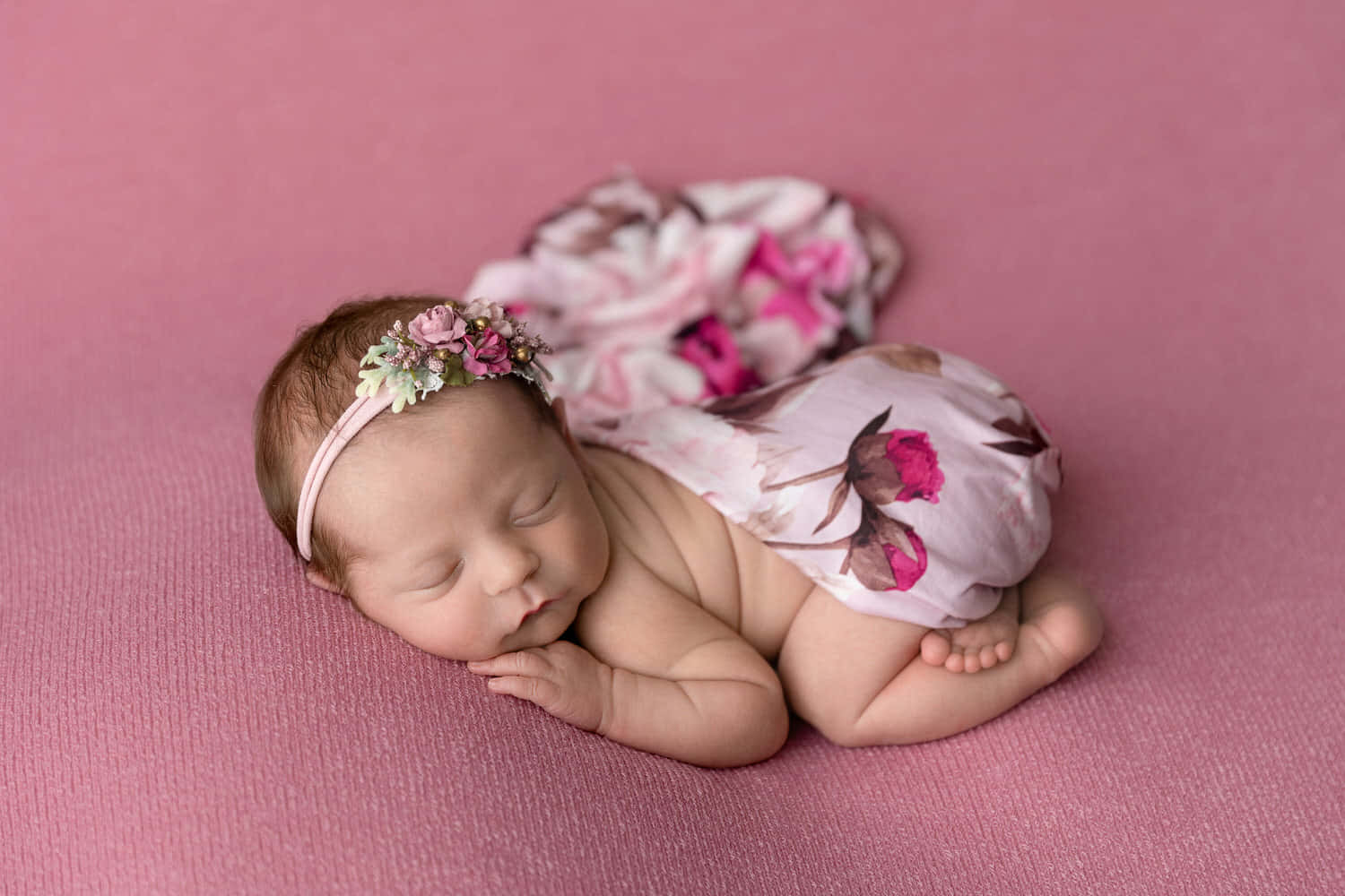 Newborn Baby 桌布 壁紙 — 一個平靜的新生嬰兒蜷縮在柔軟的粉紅色紋理表面上睡覺，戴著花卉頭帶和相配的花卉圖案衣服。