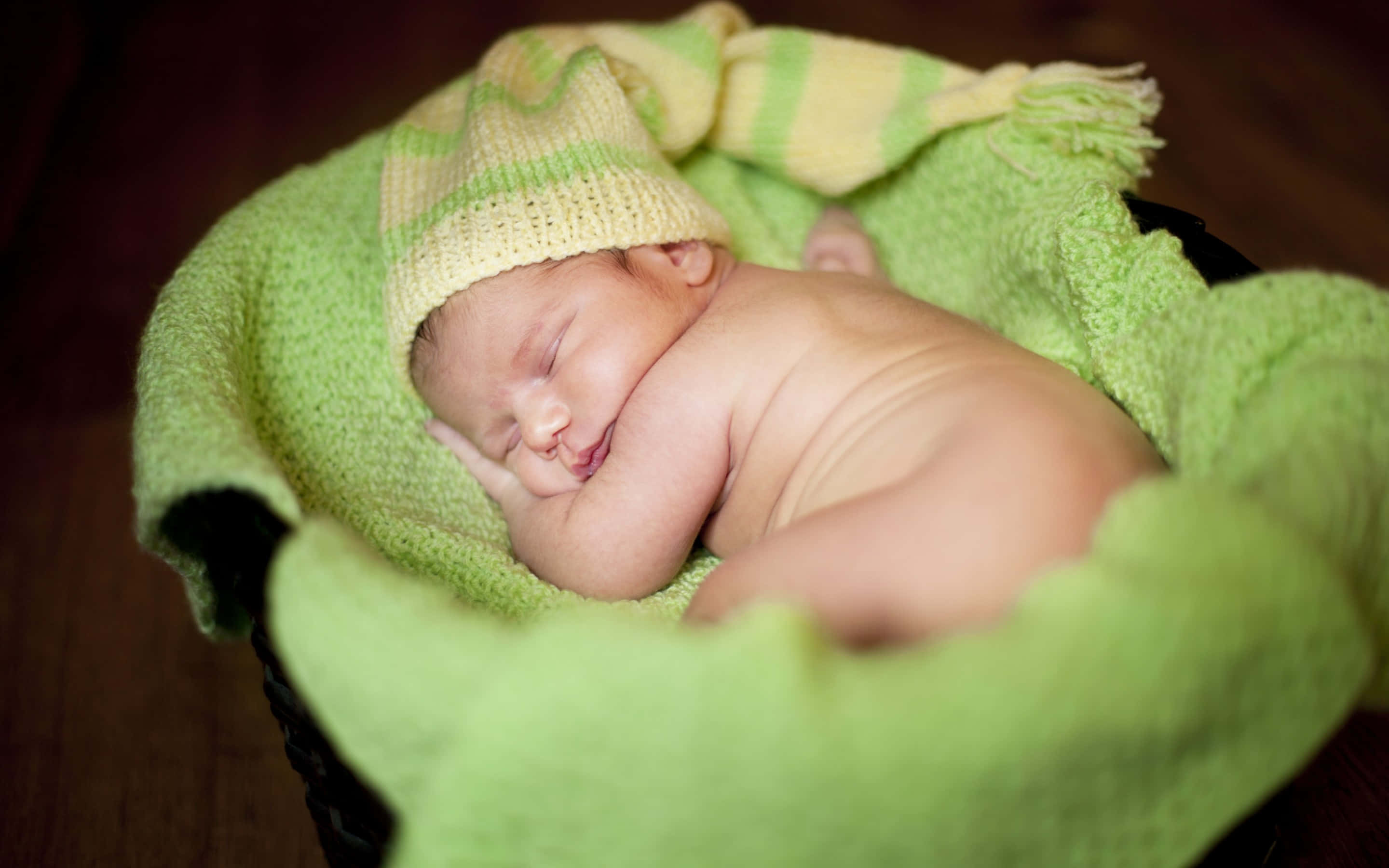 Infant 桌布 壁紙 — 一個平靜的新生嬰兒蜷縮在籃子裡睡覺，包裹在柔軟的綠色針織布料中，戴著相配的條紋帽子。