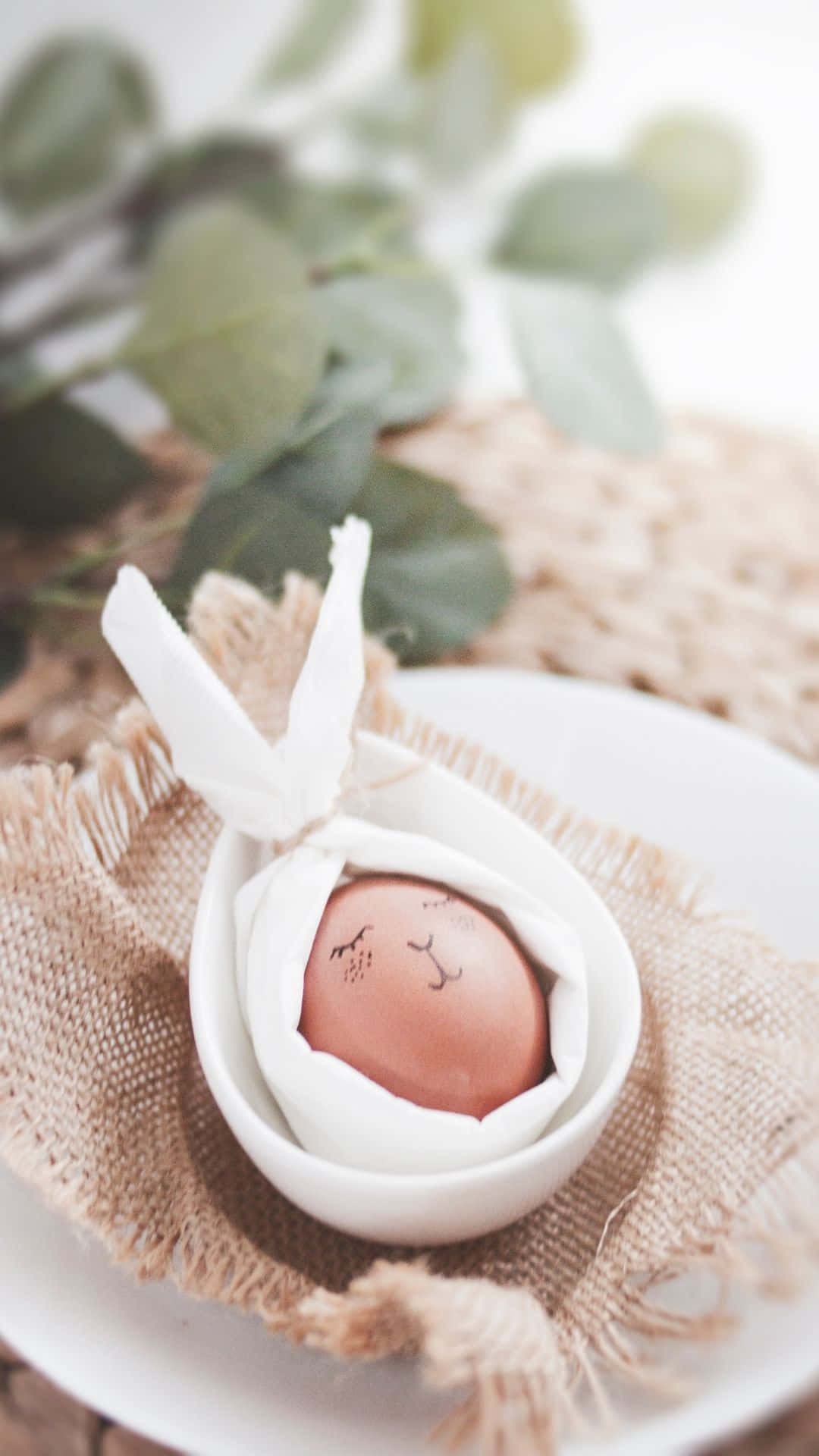 Easter Egg 桌布 壁紙 — 一個平靜的特寫，一個棕色雞蛋上畫有兔子臉，放在一個帶有兔子耳朵的白色陶瓷碗中，擱在白色盤子上的粗麻布上，背景有模糊的綠色植物。