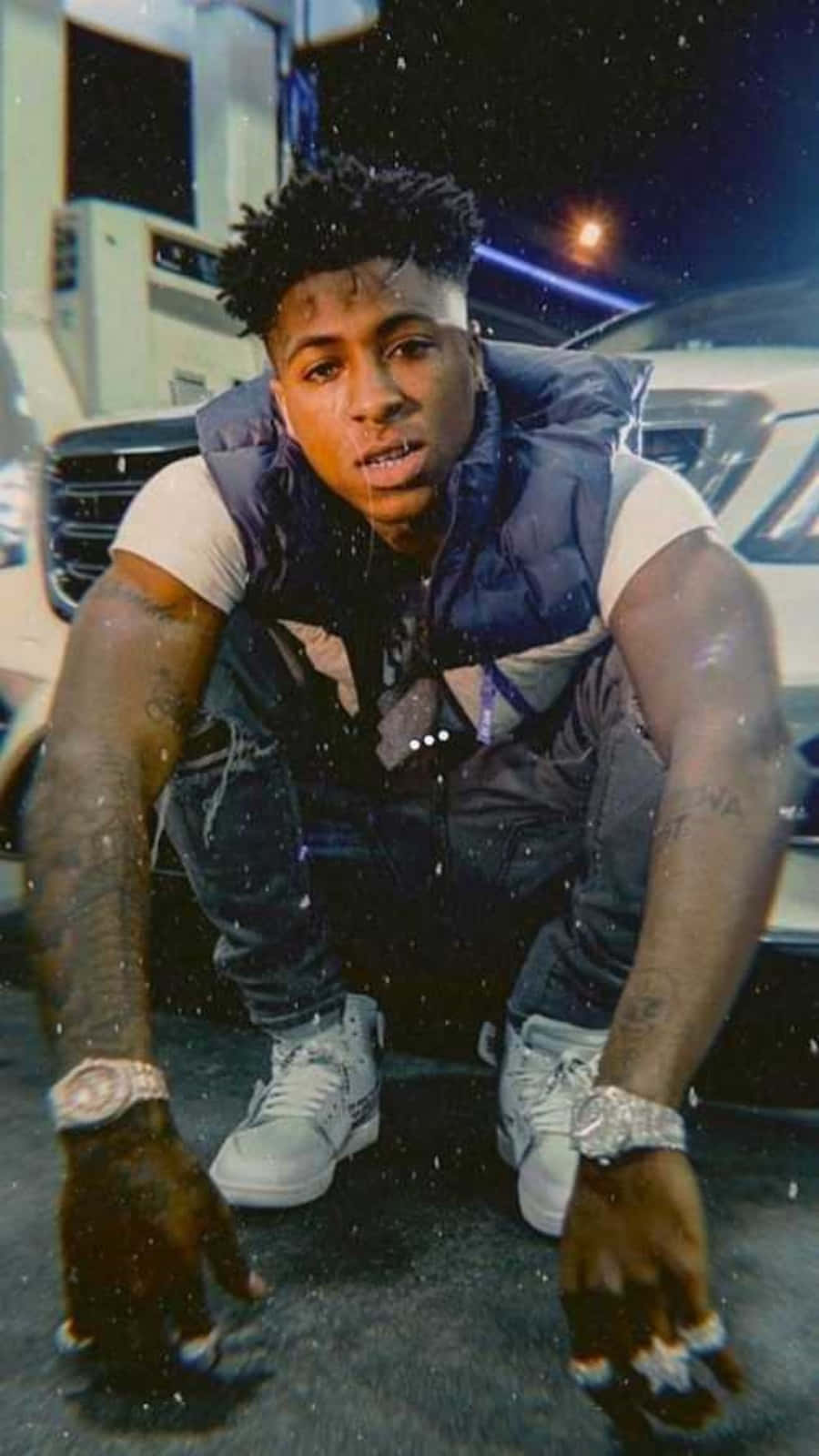 NBA YoungBoy 桌布 壁紙 — 一個年輕人在夜晚雨中自信地蹲在一輛豪華車旁邊，背景有發光的霓虹燈。
