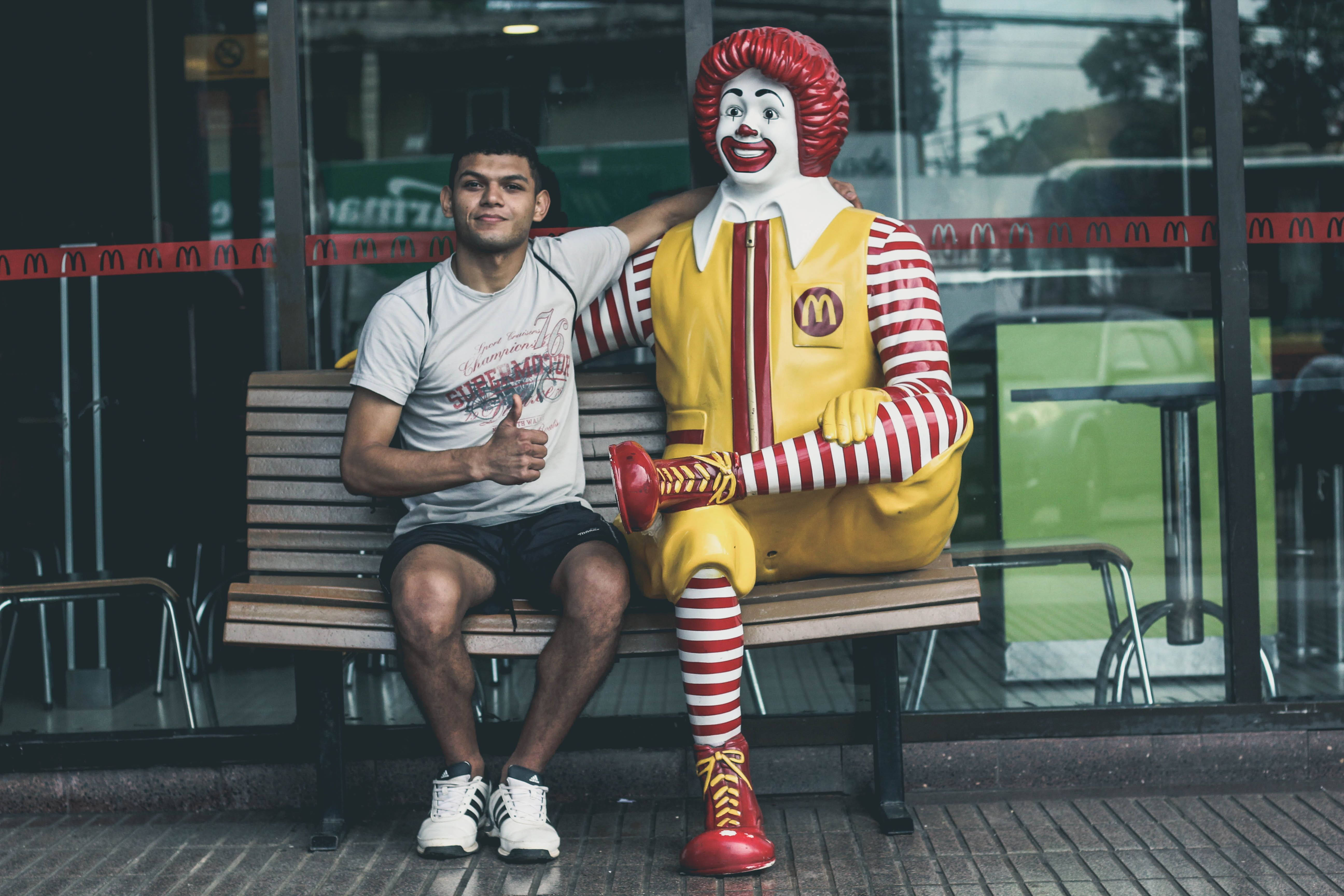 Mcdonalds 桌布 壁紙 — 一個年輕人在麥當勞外的木長椅上，坐在一個真人大小的羅納德麥當勞雕像旁邊，豎起大拇指。