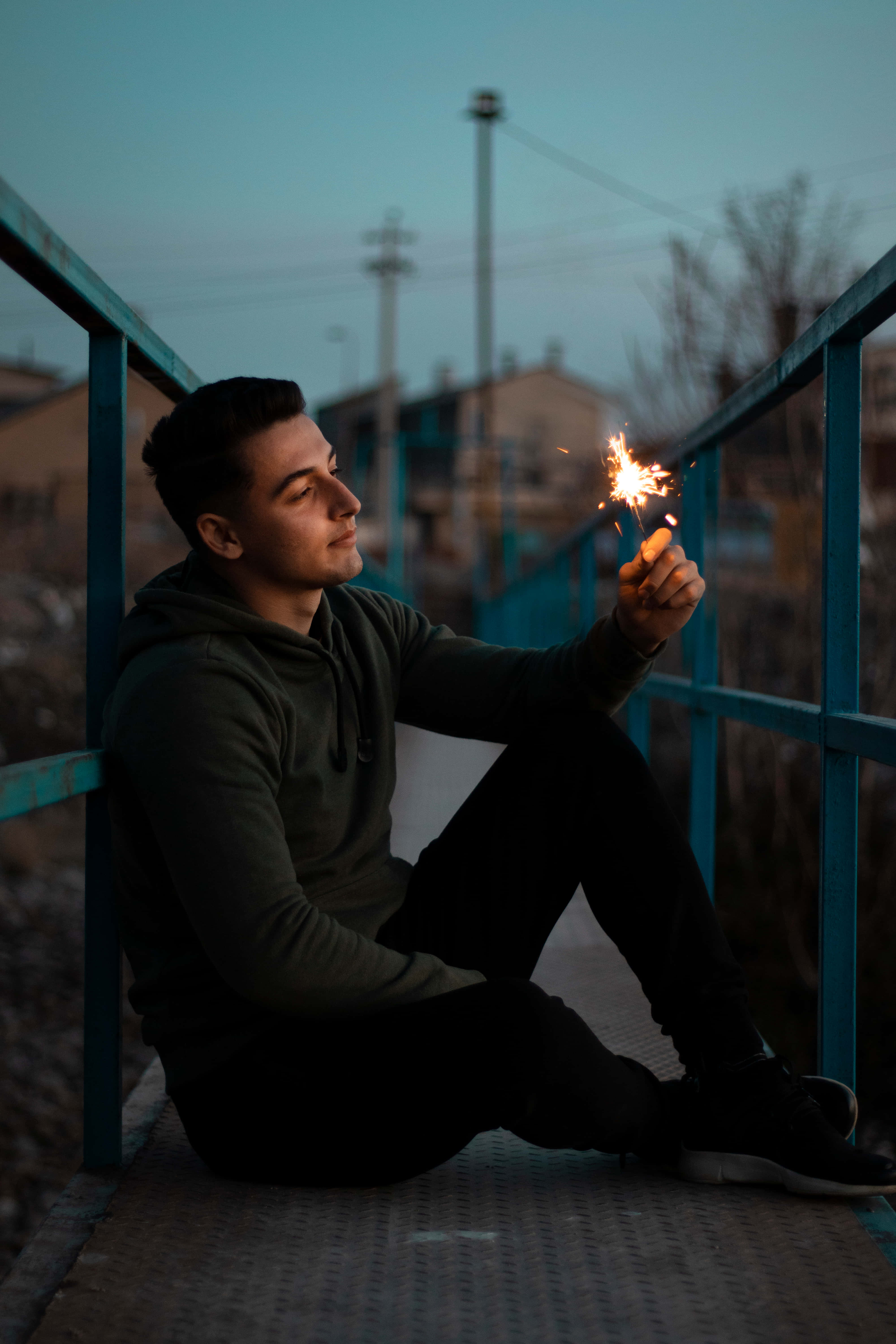 sadness 桌布 壁紙 — 一個年輕人在黃昏時坐在藍色金屬橋上，手持閃爍的煙火，他的臉被其溫暖的光芒照亮。