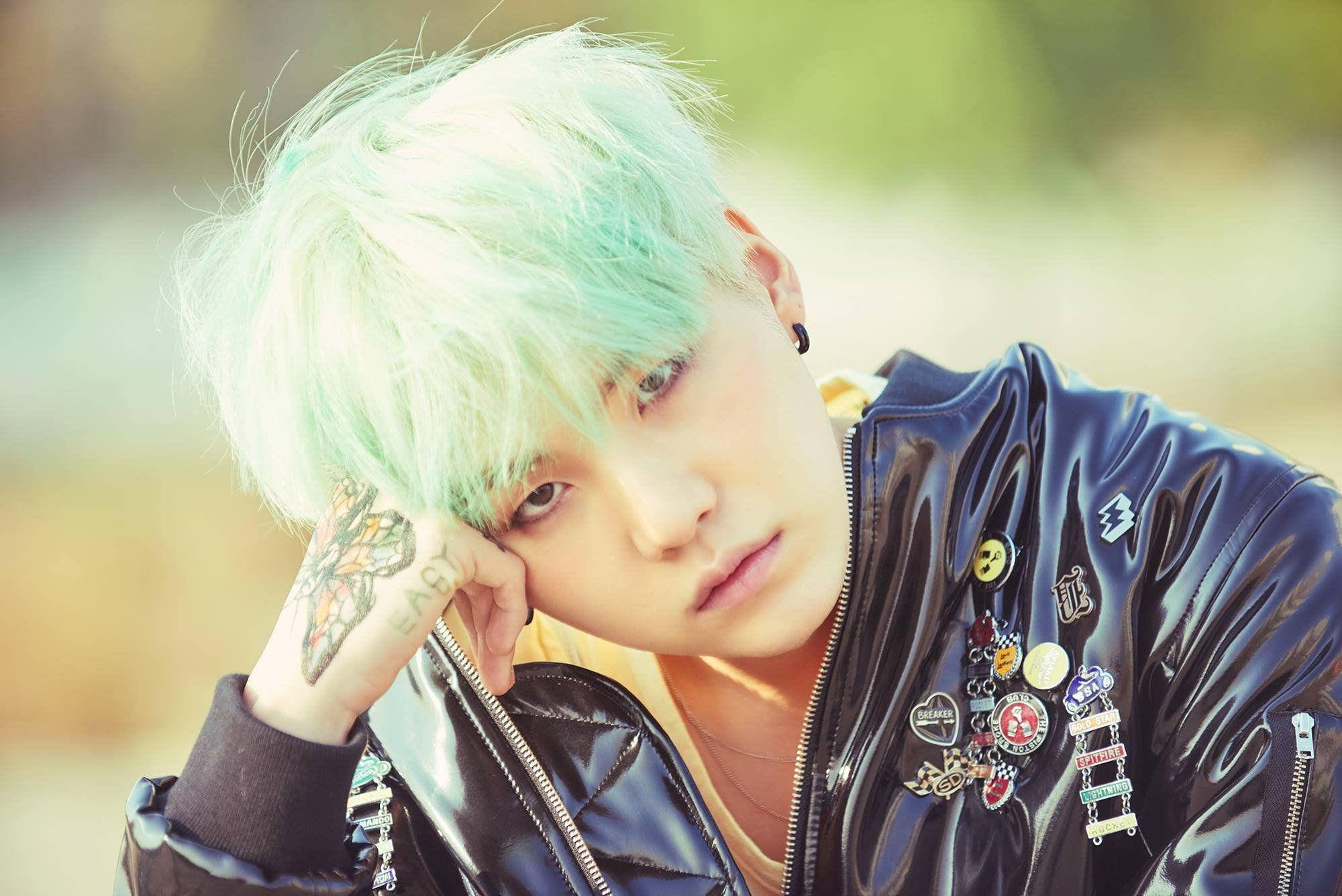 Suga Bts 桌布 壁紙 — 一個年輕人擁有薄荷綠色頭髮，將頭靠在有刺青的手上，身穿飾有彩色徽章的黑色外套。