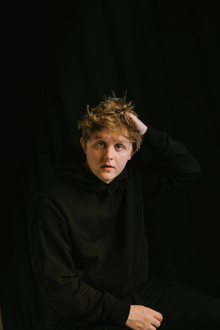 Lewis Capaldi 桌布 壁紙 — 一個年輕人有凌亂的金髮，坐在黑色背景前，手放在頭上，穿著黑色連帽衫，在戲劇性的工作室燈光下。