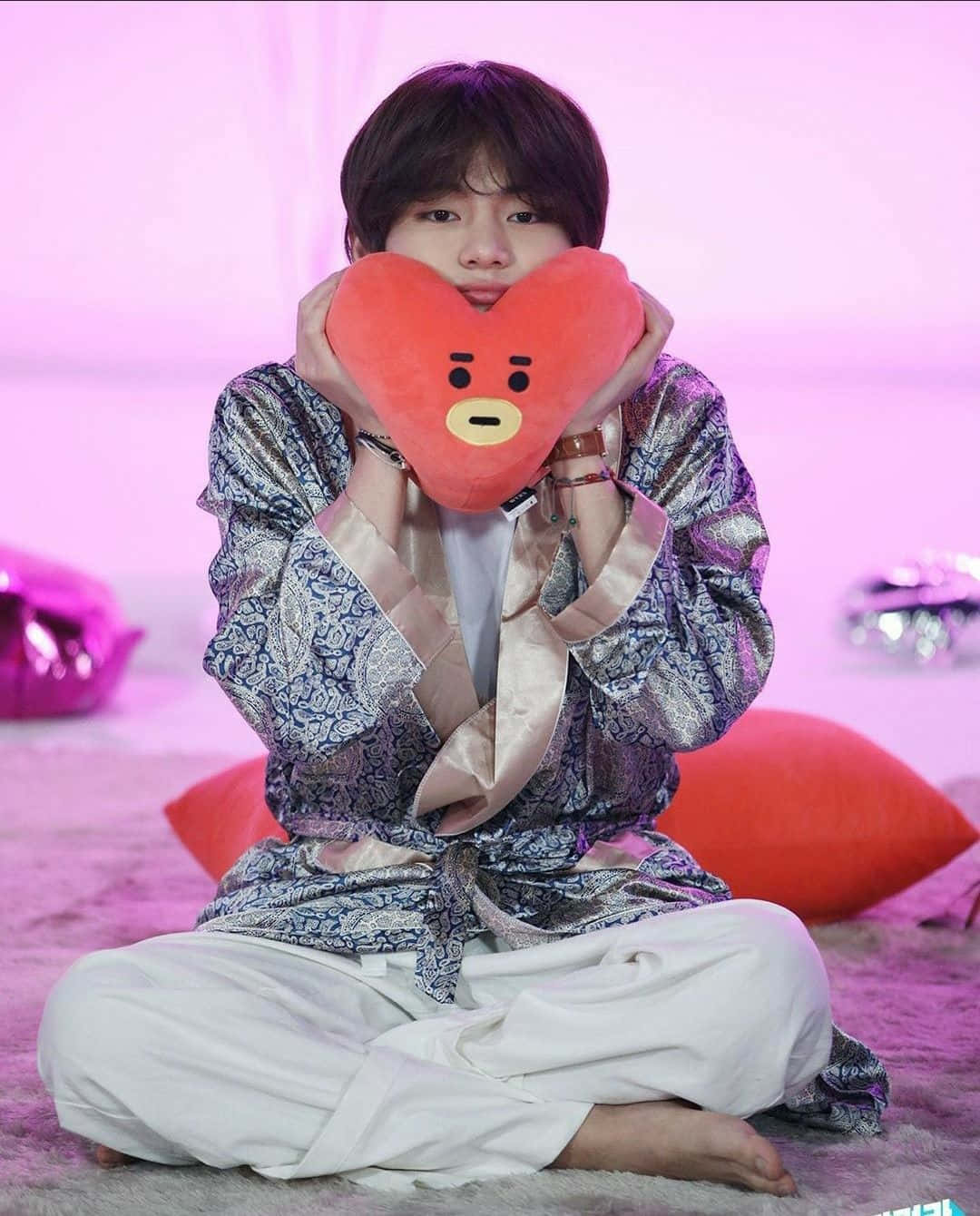 Taehyung Cute 桌布 壁紙 — 一個年輕人盤腿而坐，在柔和的粉紅色背景前抱著一個帶有卡通臉的大紅色心形毛絨玩具。