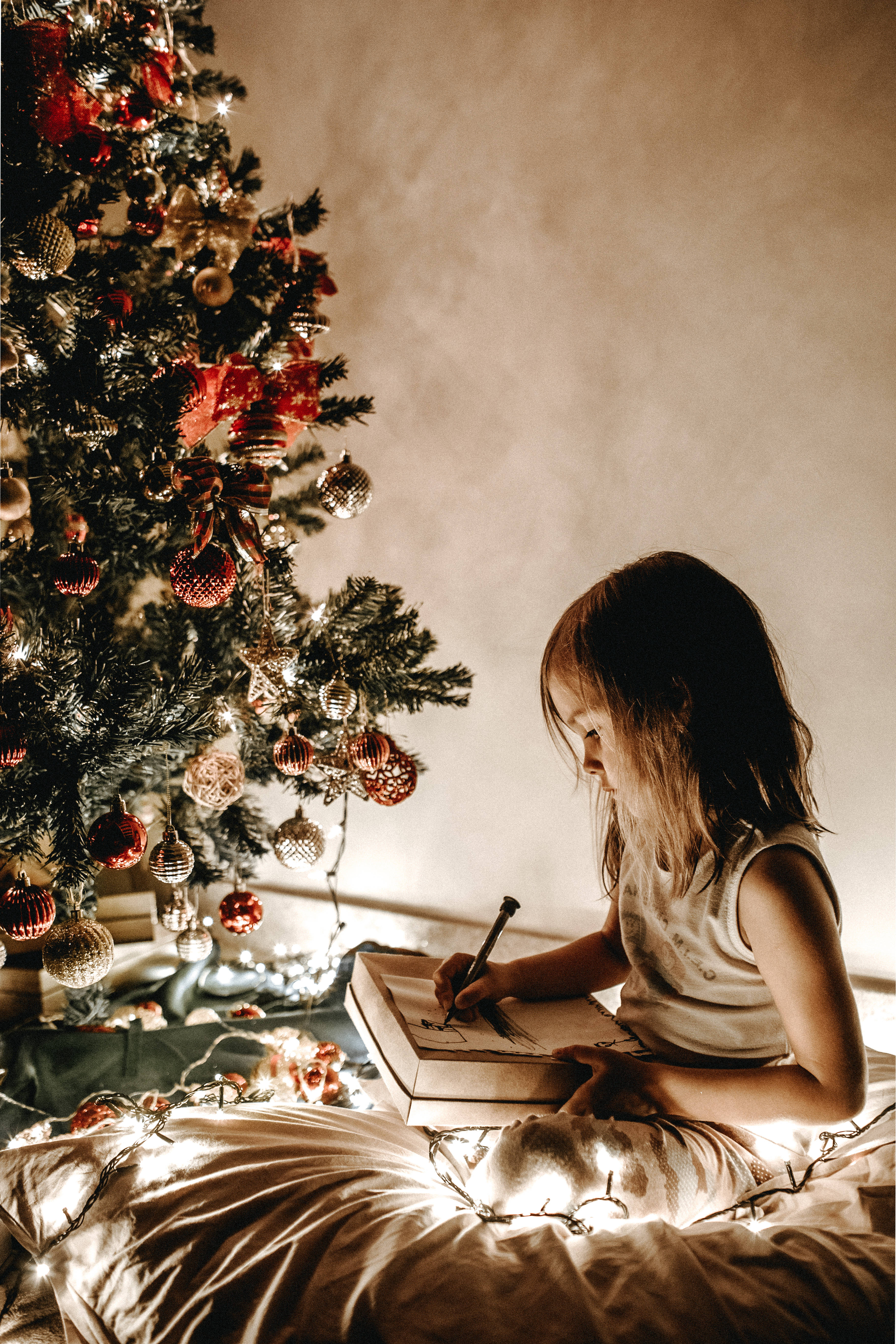 Santa Claus 桌布 壁紙 — 一個年輕女孩坐在床上，周圍環繞著閃爍的童話燈光和裝飾好的聖誕樹，同時在筆記本上畫畫。