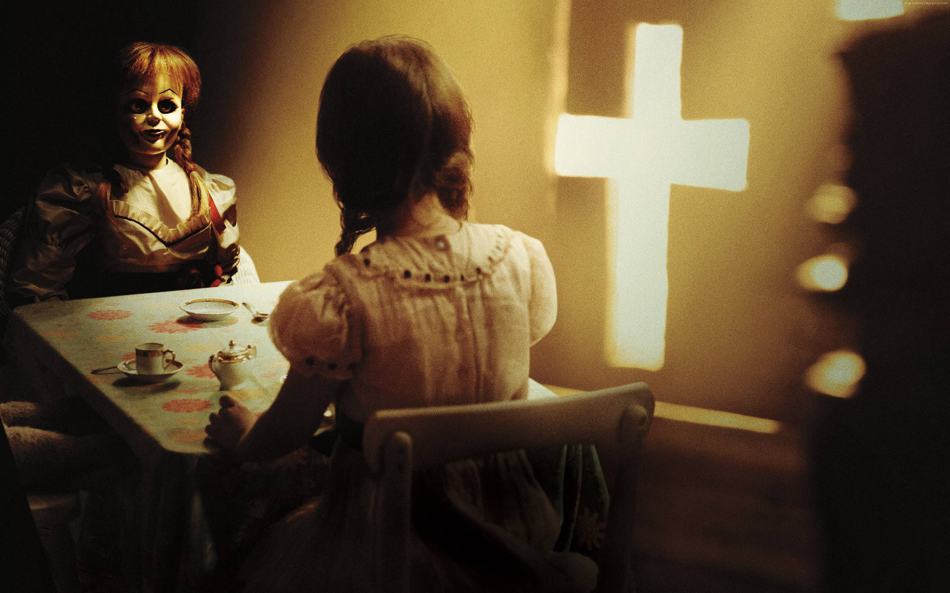 Annabelle 桌布 壁紙 — 一個年輕女孩坐在桌子前，面對一個畫著臉的可怕娃娃，在溫暖昏暗的燈光下，身後牆上有一個發光的十字架。