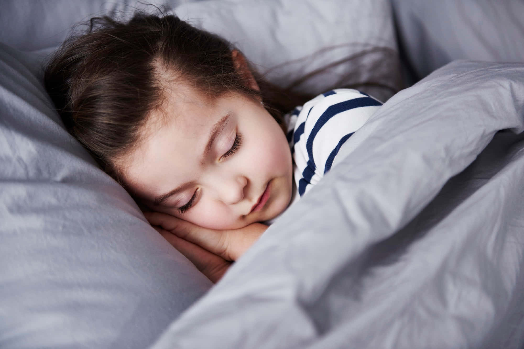 Sleep 桌布 壁紙 — 一個年輕女孩將頭枕在手上，依偎在柔軟的灰色寢具中，安詳地睡著。