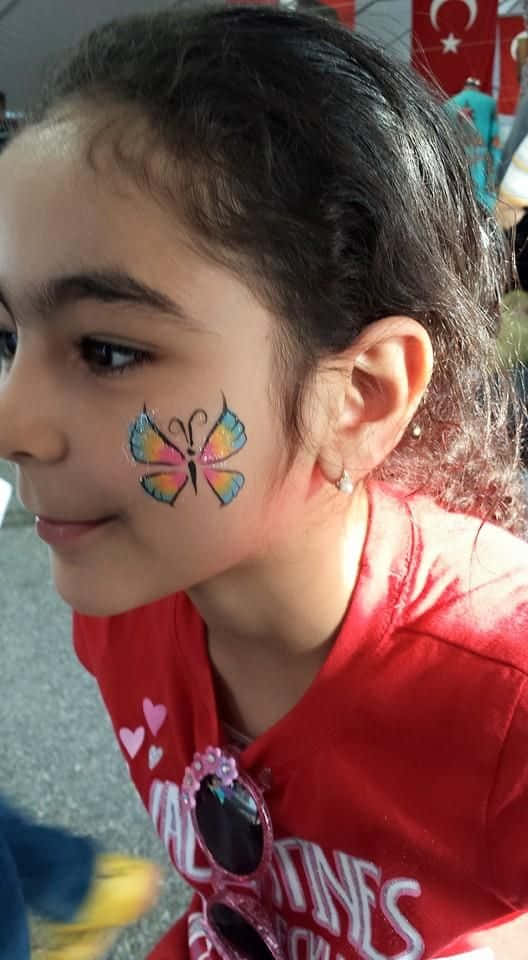 Butterfly Face Painting 桌布 壁紙 — 一個年輕女孩微笑，臉頰附近有色彩繽紛的蝴蝶臉部彩繪，穿著紅色襯衫。