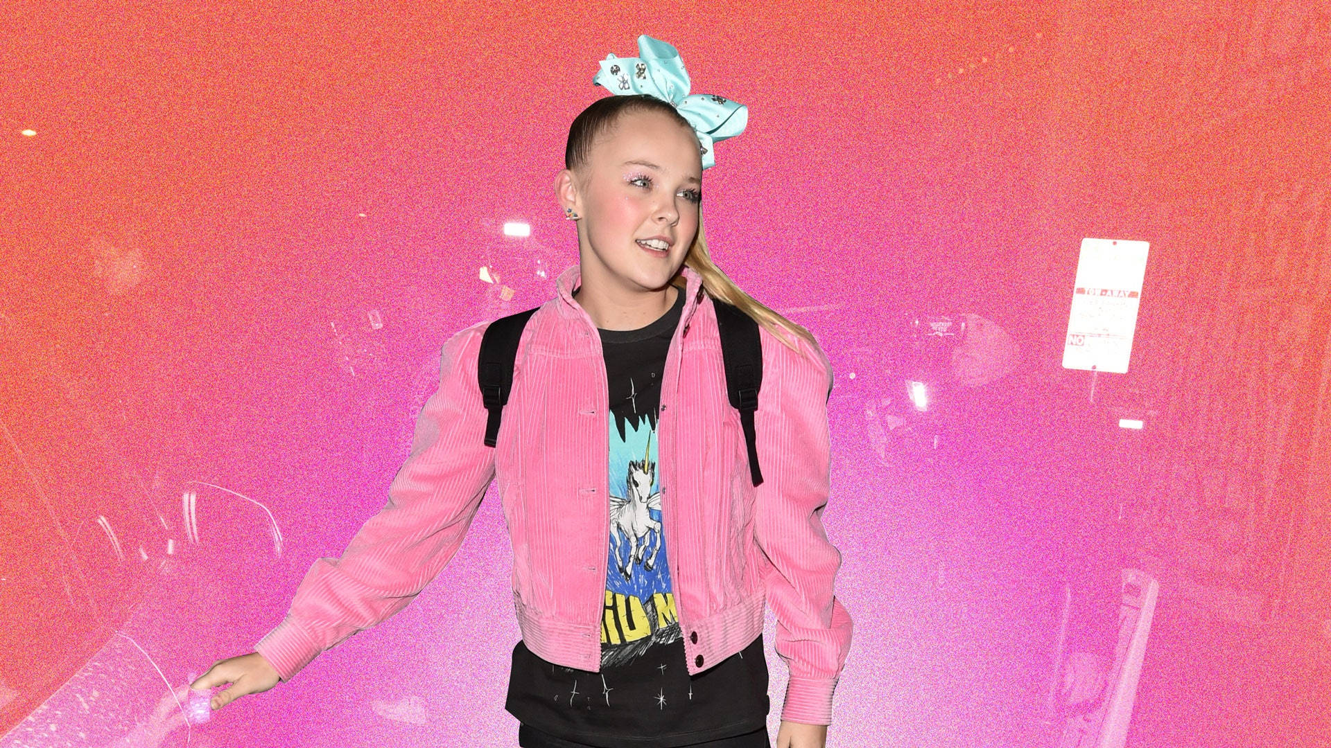 JoJo Siwa 桌布 壁紙 — 一個年輕女孩戴著一個大藍色蝴蝶結，穿著粉紅色外套，在充滿活力的粉紅色和橙色漸變牆前微笑。