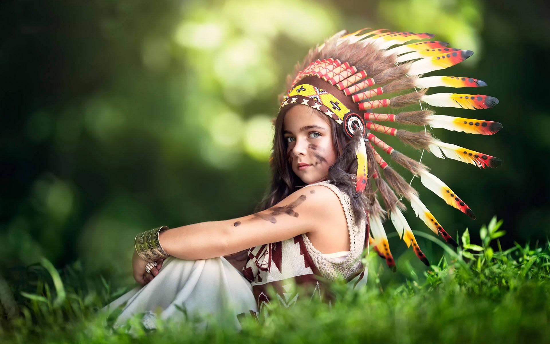 girls 桌布 壁紙 — 一個年輕女孩戴著色彩繽紛的羽毛頭飾，優雅地坐在茂密的綠草中，柔和的陽光從她身後的樹木間透射進來。