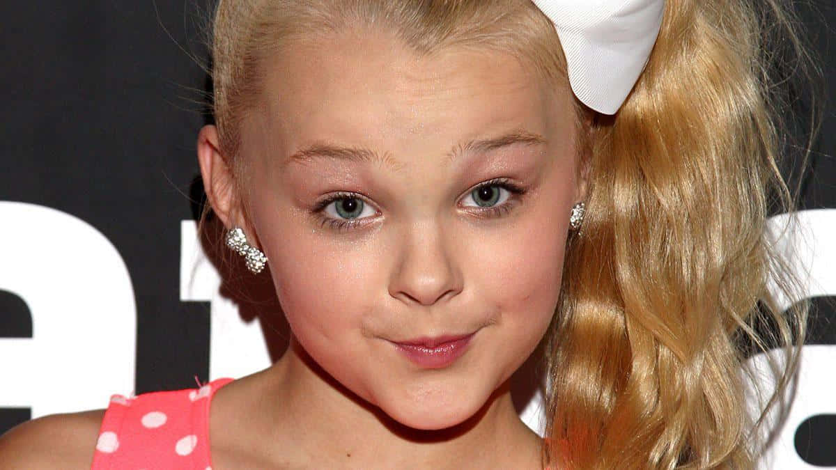 JoJo Siwa 桌布 壁紙 — 一個年輕女孩的頭像特寫，金髮紮成高馬尾，飾有白色蝴蝶結，身穿粉紅色圓點上衣和閃亮耳環。