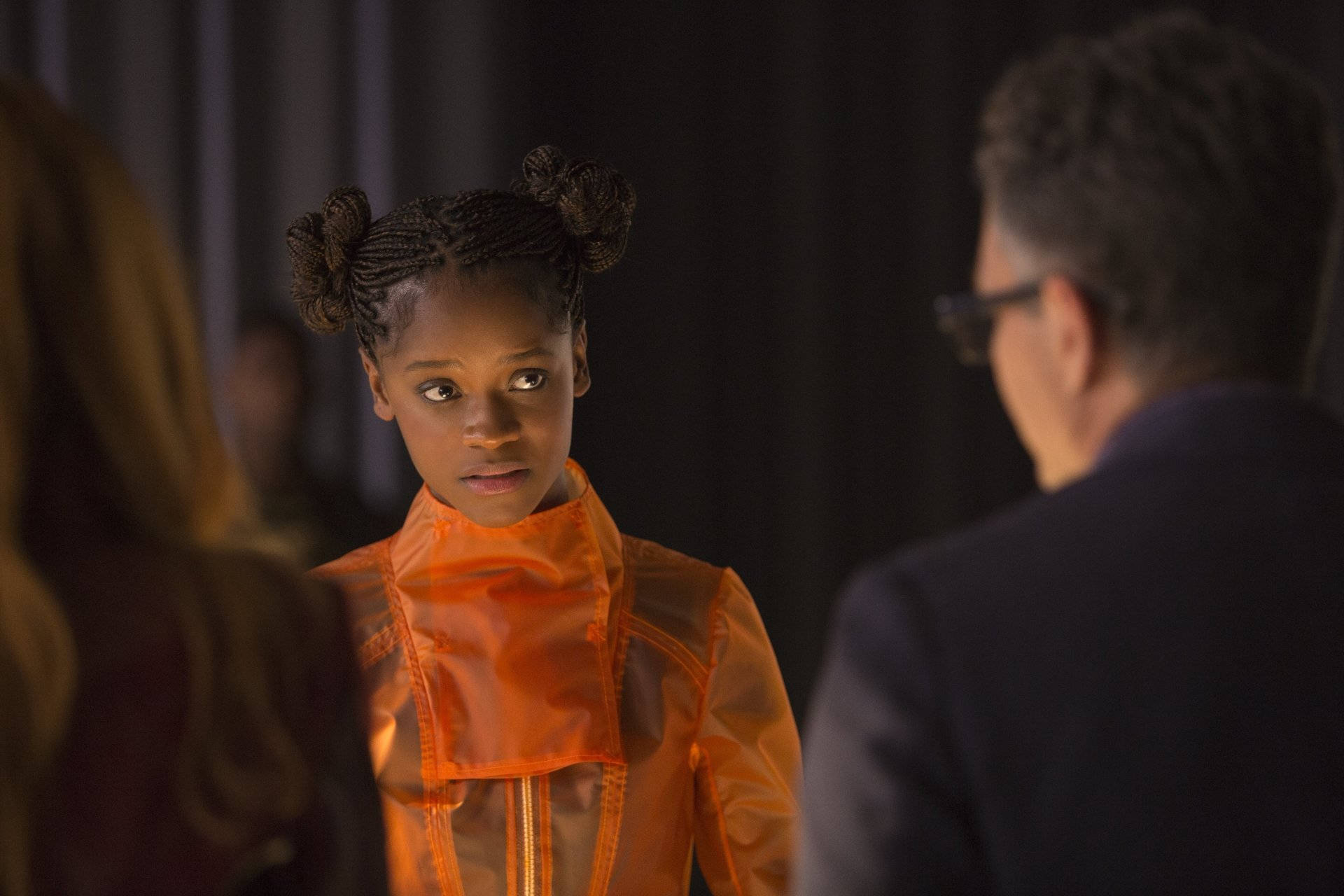 Shuri 桌布 壁紙 — 一個年輕女孩穿著亮橙色半透明服裝，編著辮子，在昏暗的房間裡專注地看著一個成年人。
