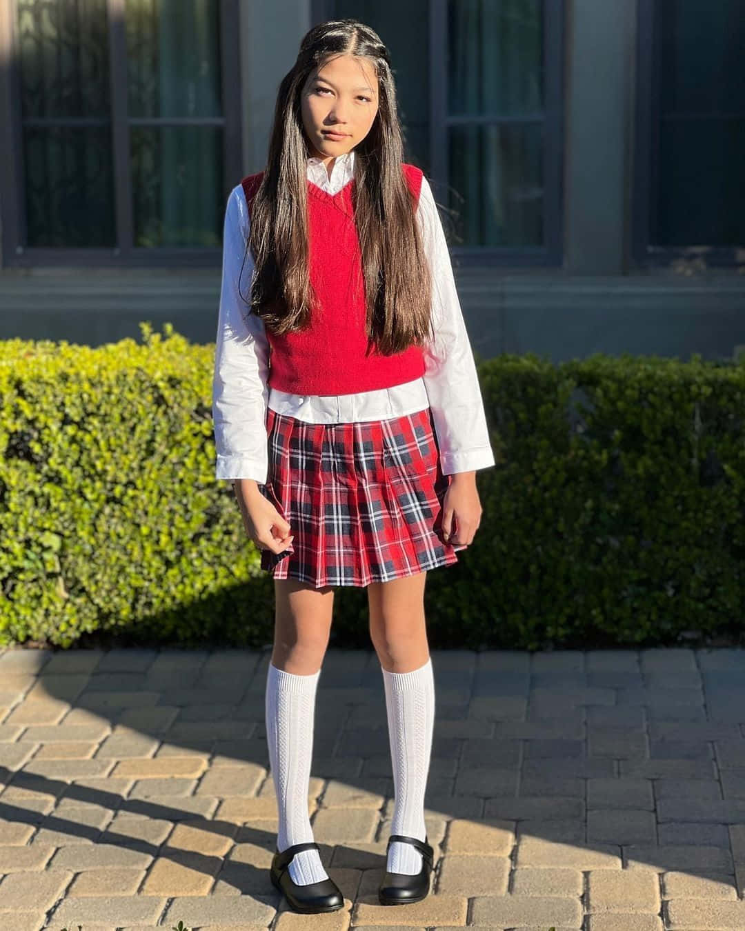美學 桌布 壁紙 — 一個年輕女孩穿著紅色背心、格子裙和白色襪子，自信地站在陽光普照的磚砌小徑上，身後有綠色樹籬。