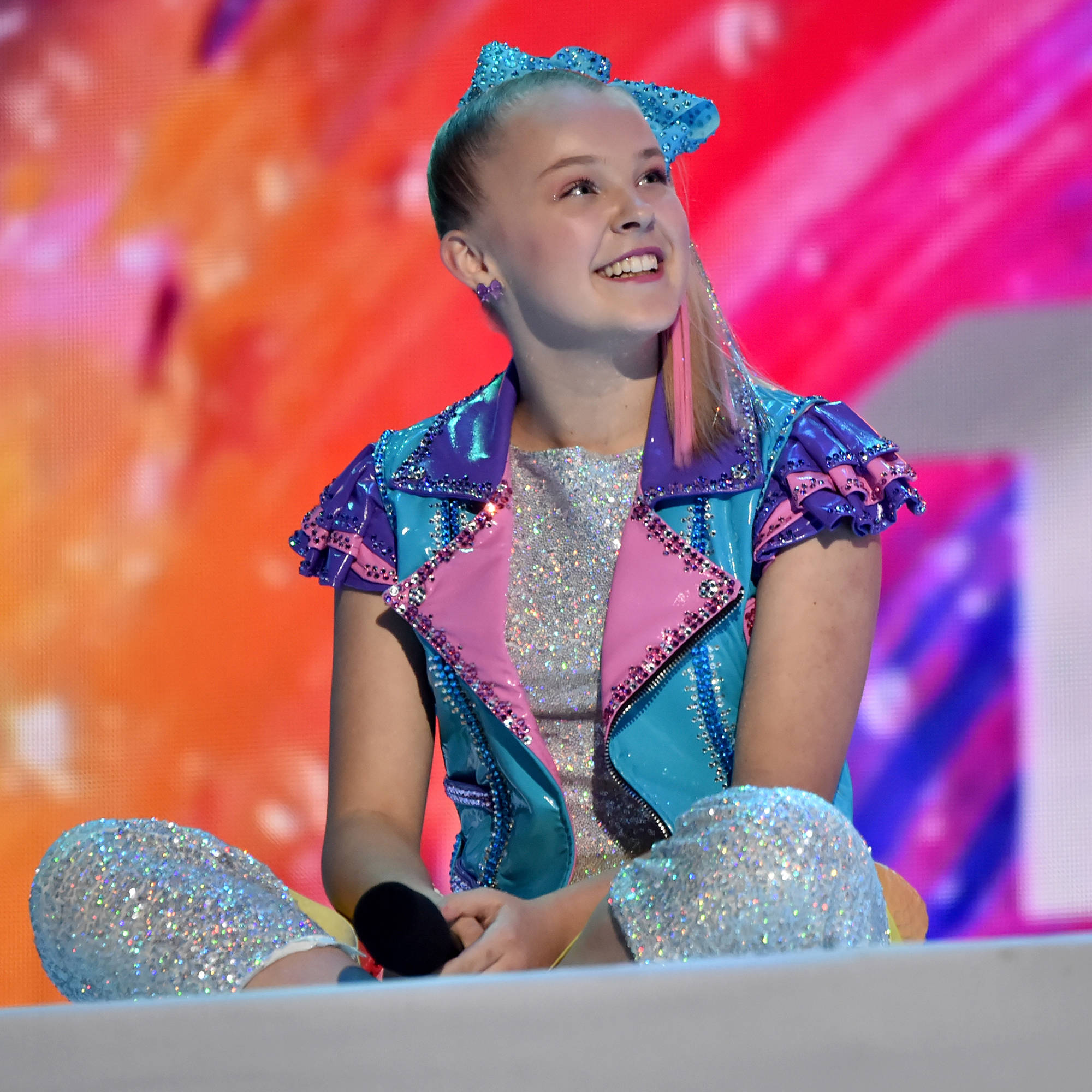 JoJo Siwa 桌布 壁紙 — 一個年輕女孩穿著閃閃發光的藍色、粉紅色和銀色服裝，坐在色彩繽紛的抽象背景舞台上，露出燦爛的笑容。