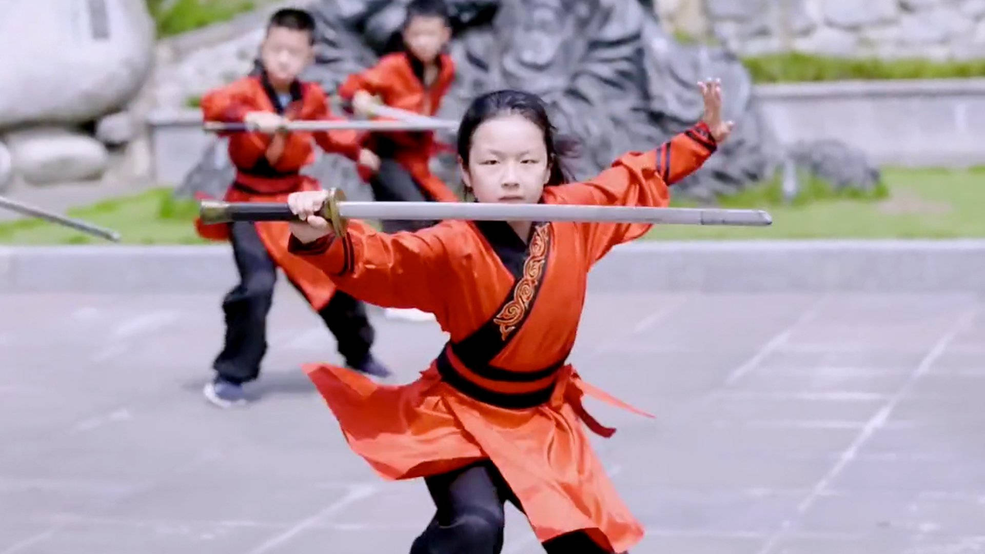小孩動圖 桌布 壁紙 — 一個年輕女孩穿著鮮豔的橙色武術制服，專注地揮舞著劍，而兩個男孩在她身後的一個石頭庭院裡訓練。