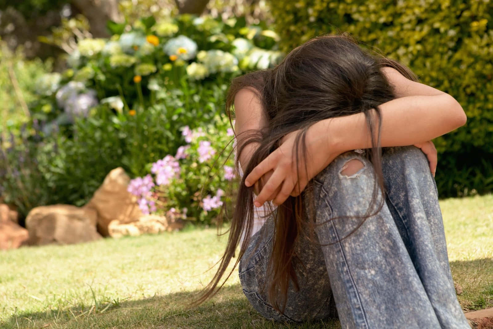 Depressed 桌布 壁紙 — 一個年輕女孩蜷縮在陽光花園的草地上，頭埋在手臂中，周圍環繞著綠色植物和紫色花朵。