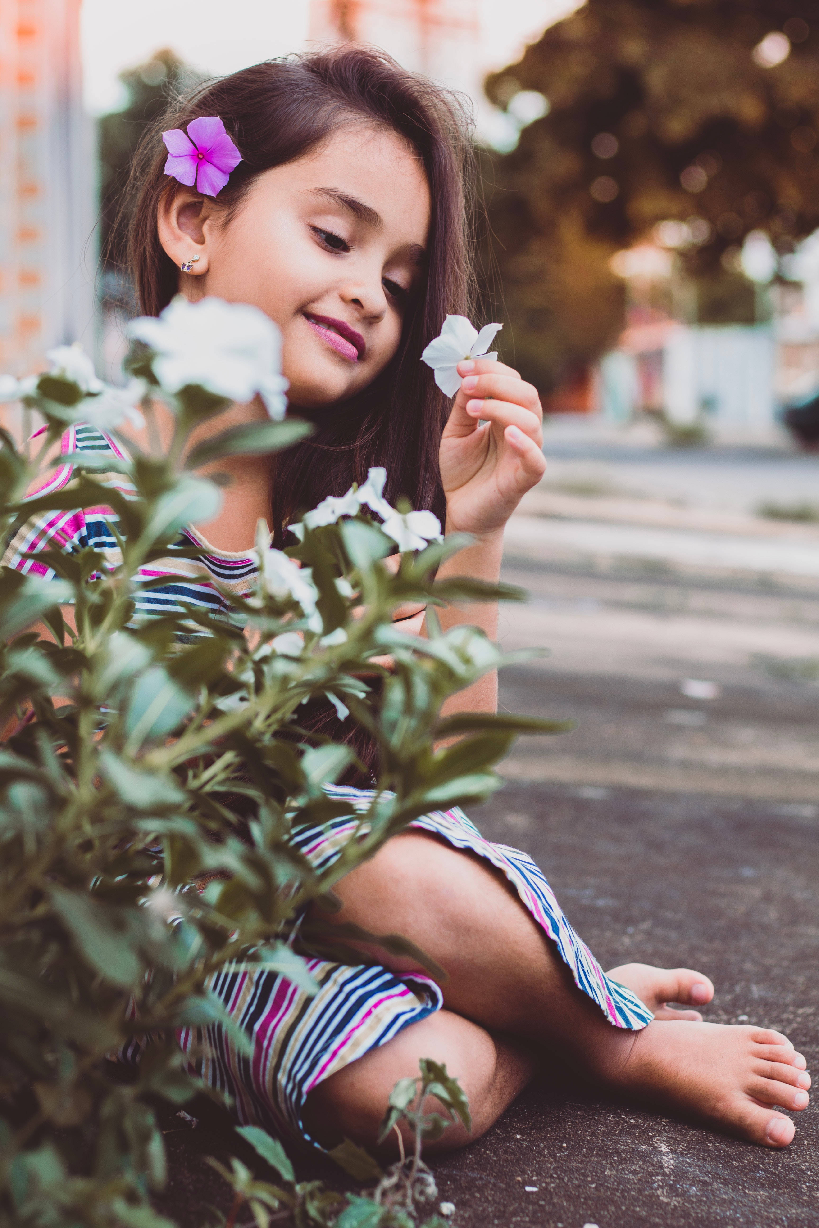 小孩動圖 桌布 壁紙 — 一個年輕女孩頭髮上插著一朵紫色花，赤腳坐在人行道上，輕輕拿著一朵白色花，溫柔地微笑。