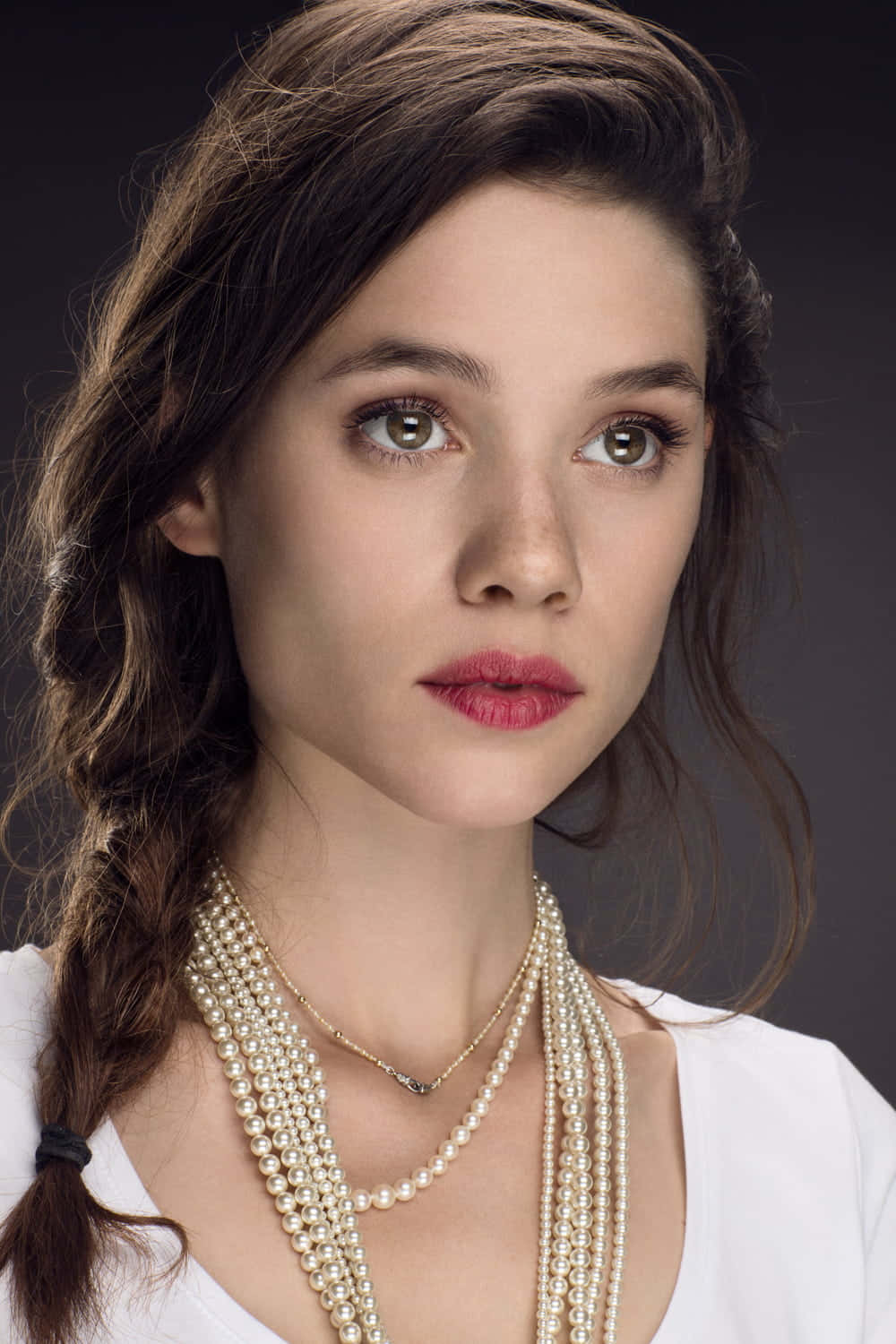 Astrid Berges Frisbey 桌布 壁紙 — 一個年輕女性的特寫肖像，棕色頭髮編成辮子，戴著多層珍珠項鍊和紅色唇膏，背景為深色。