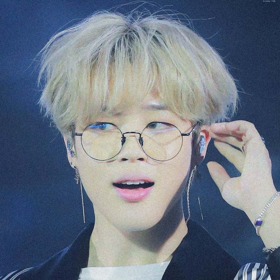 Jimin Aesthetic 桌布 壁紙 — 一個年輕男子，一頭凌亂的金髮，戴著圓框眼鏡，在舞台燈光下調整耳機的特寫。
