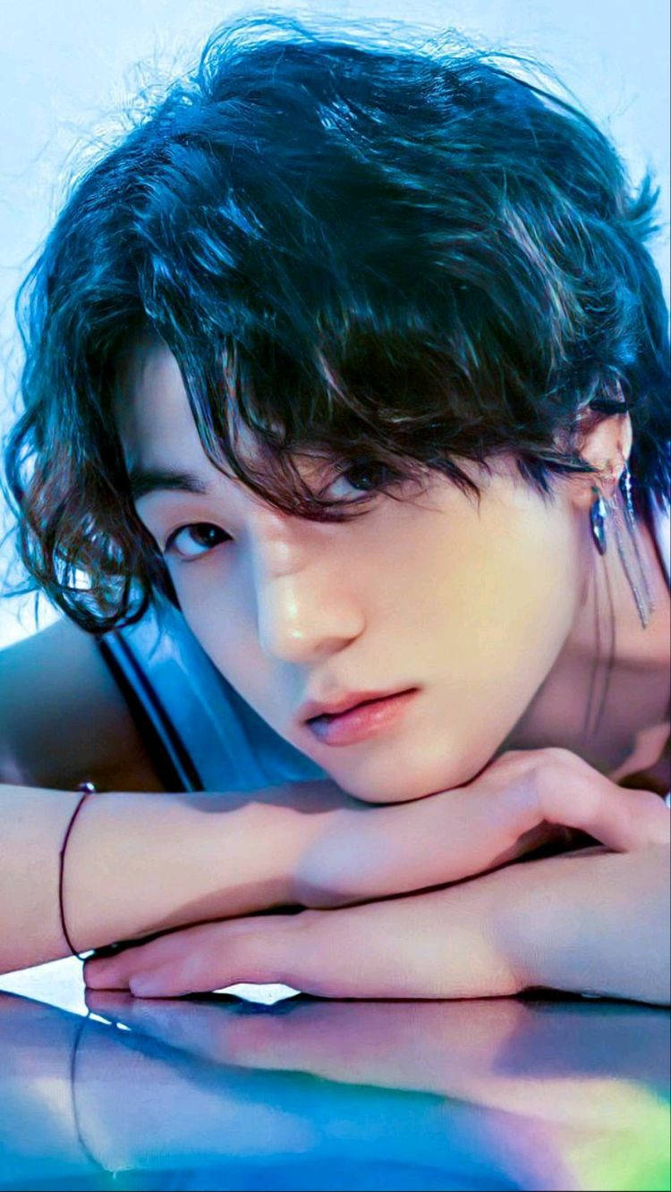 Jungkook 桌布 壁紙 — 一個年輕男子，一頭凌亂的黑髮，在冰冷的藍色燈光下，將下巴擱在交疊的雙手上，靠著反光表面。