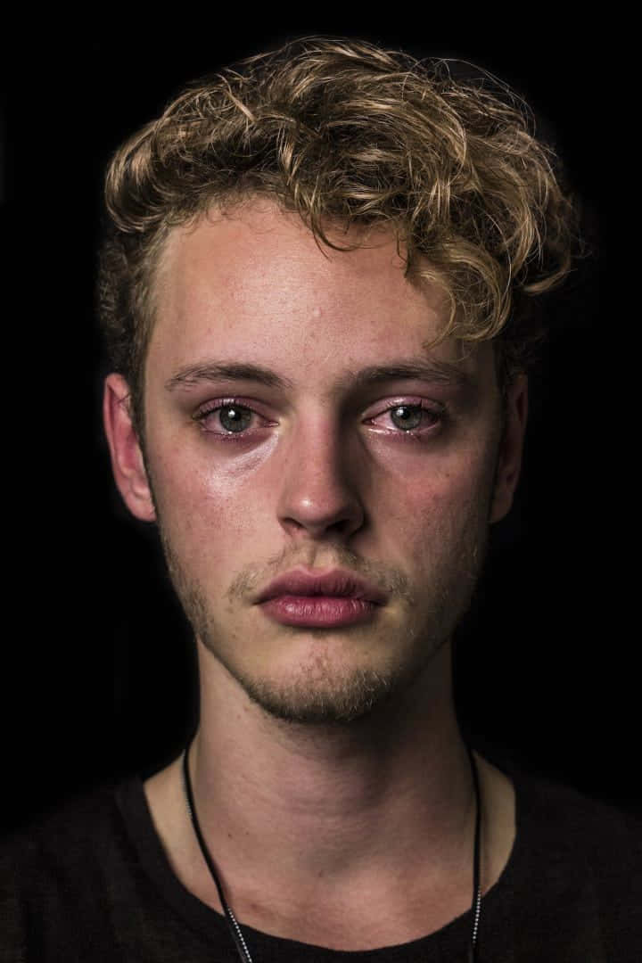 Alone 桌布 壁紙 — 一個年輕男子，有著捲曲的金色頭髮和疲憊的眼睛，在黑色背景下強烈地凝視著。