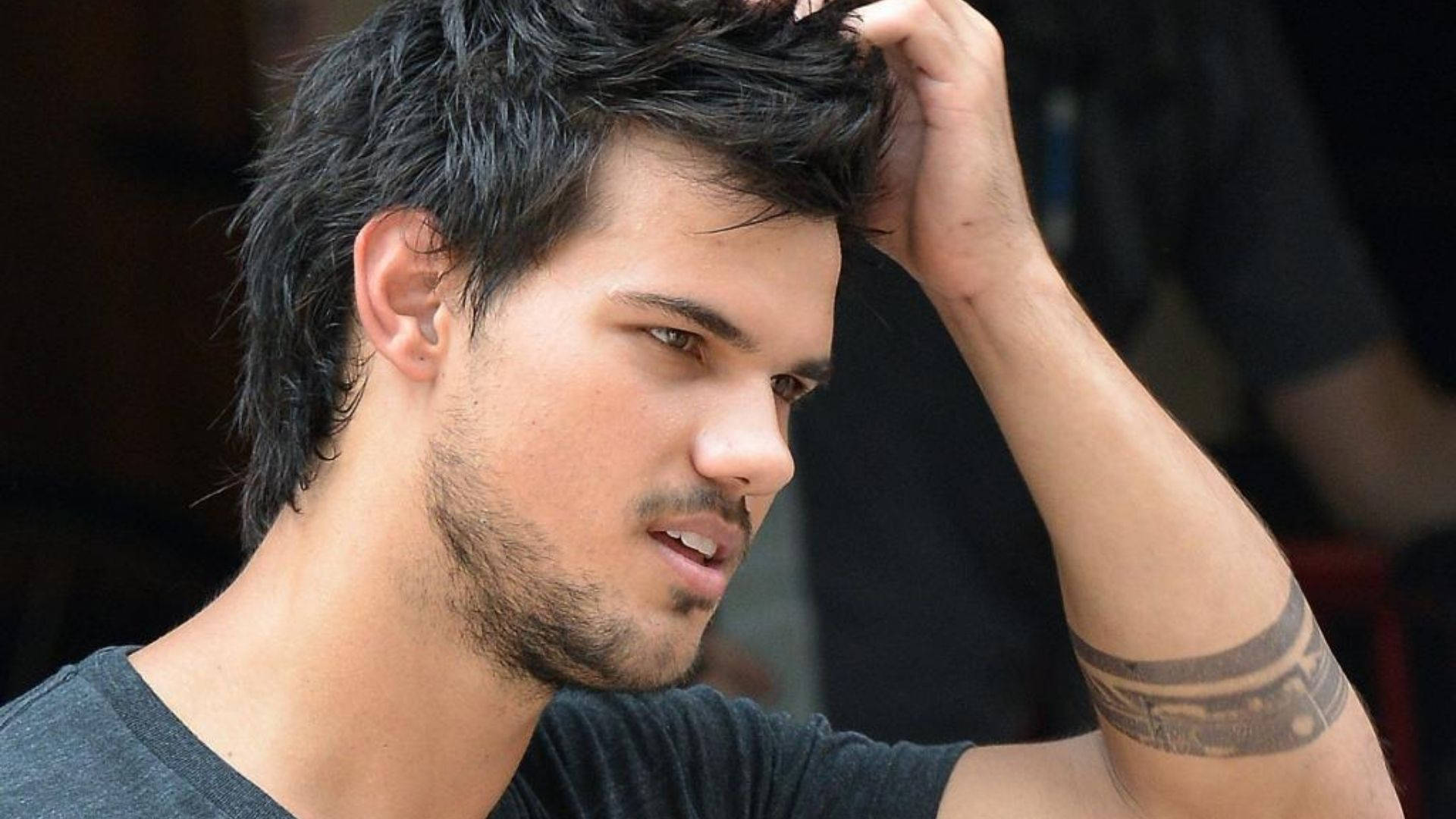 Taylor Lautner 桌布 壁紙 — 一個年輕男子，深色凌亂的頭髮和有刺青的手臂，一邊用手撥弄頭髮，一邊望向旁邊。