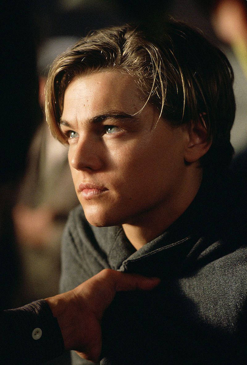 Dicaprio 桌布 壁紙 — 一個年輕男子，頭髮蓬亂，藍眼睛深邃，在溫暖的電影燈光下側臉沉思。