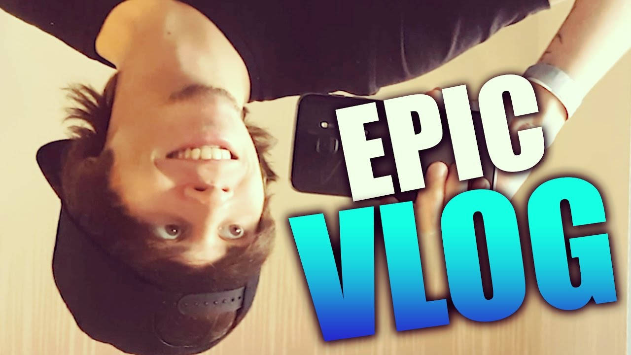 YouTube 桌布 壁紙 — 一個年輕男子倒轉咧嘴笑，同時手持帶有粗體「EPIC VLOG」文字疊加的手機。