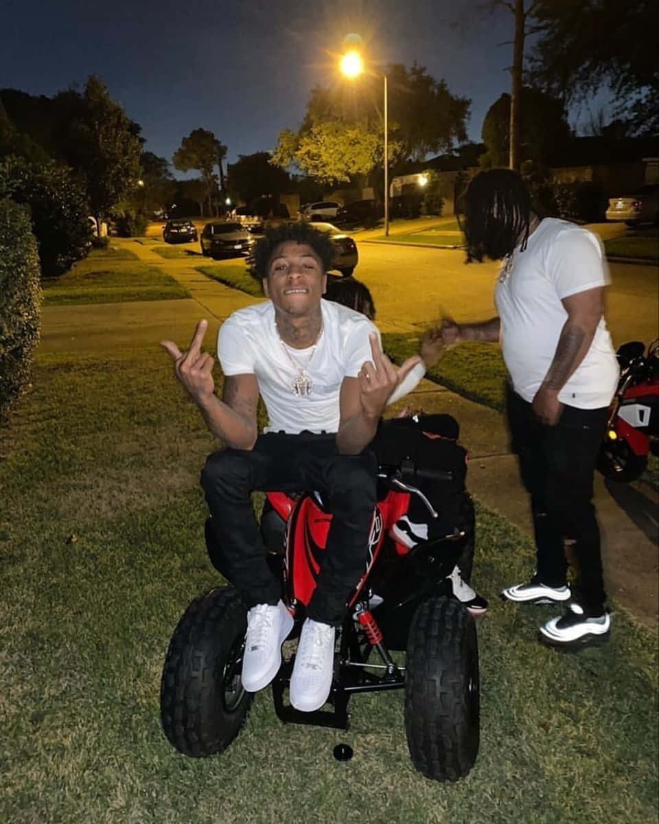 Youngboy 桌布 壁紙 — 一個年輕男子咧嘴笑著比出和平手勢，坐在紅黑相間的迷你四輪車上，夜晚在街燈下的草地上。