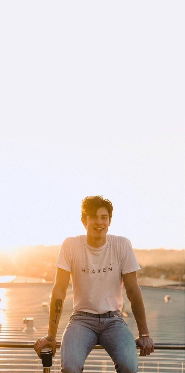 Shawn 桌布 壁紙 — 一個年輕男子坐在天台欄杆上，溫暖地微笑，金色的夕陽灑在他和遠處的城市景觀上。
