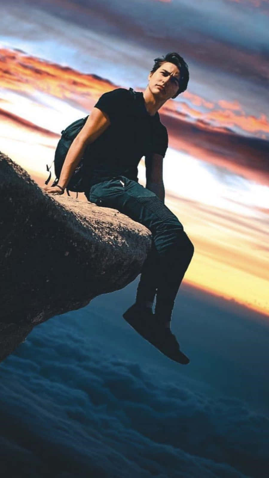 Sam And Colby 桌布 壁紙 — 一個年輕男子坐在懸崖邊，俯瞰日落時分的雲海，天空呈現火熱的橙色和藍色。