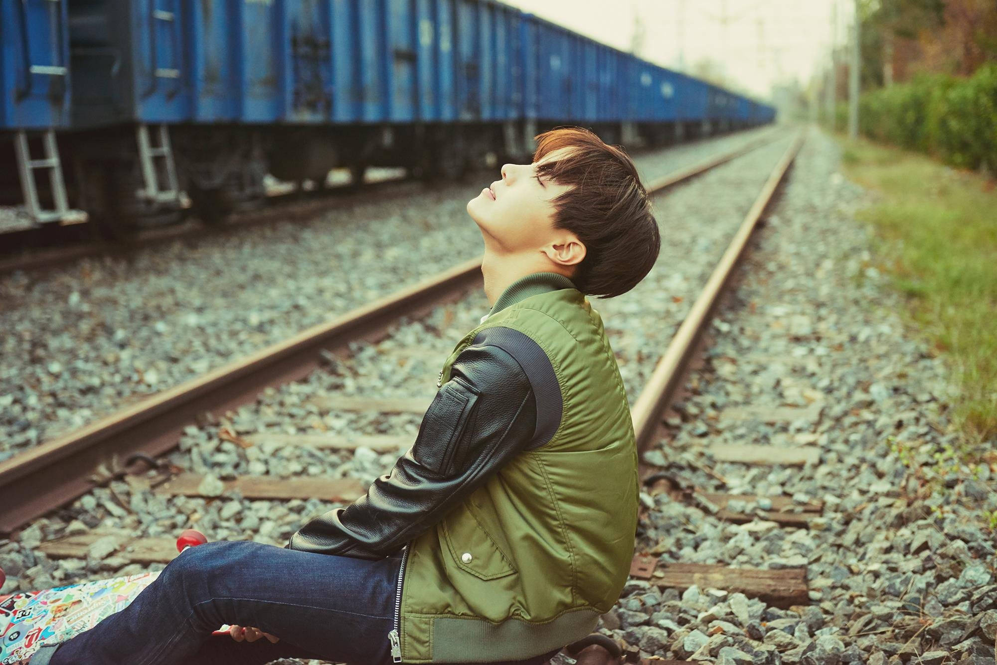 j-hope 桌布 壁紙 — 一個年輕男子坐在火車軌道上，頭向後傾，仰望旁邊一列長長的藍色貨運火車。