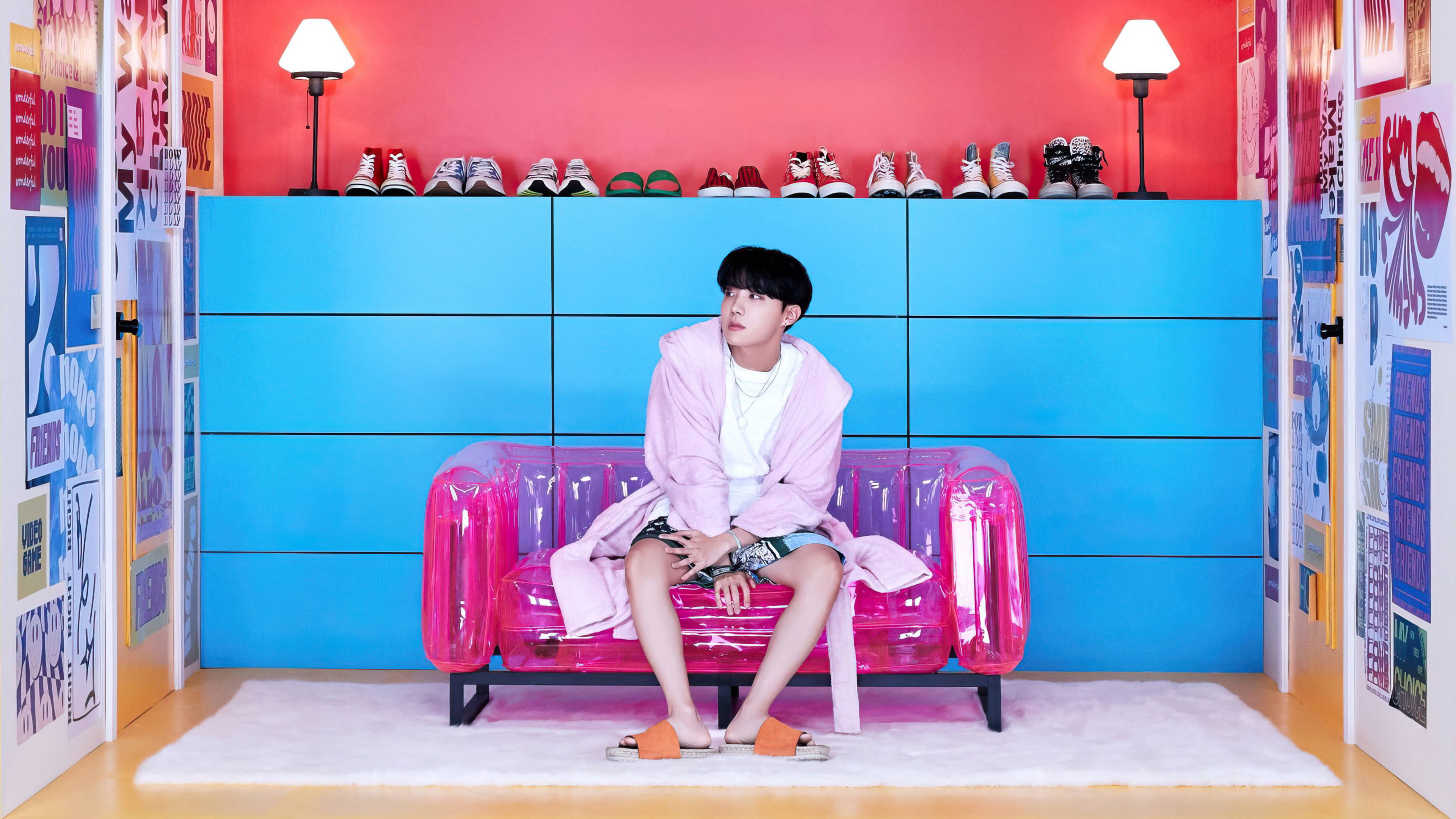 j-hope 桌布 壁紙 — 一個年輕男子坐在粉紅色充氣沙發上，房間充滿活力，藍色牆壁和彩色海報。