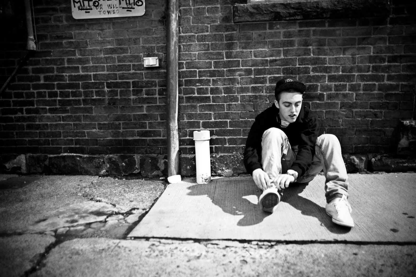Mac Miller 桌布 壁紙 — 一個年輕男子坐在裂開的人行道上，背靠磚牆繫鞋帶，呈現鮮明的黑白對比。
