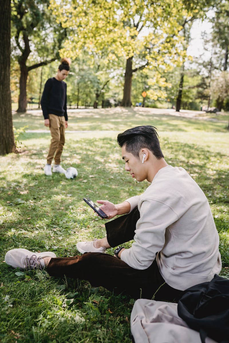 足球 桌布 壁紙 — 一個年輕男子坐在陽光明媚的公園草地上，專注於他的手機，而另一個男子則拿著足球站在附近。