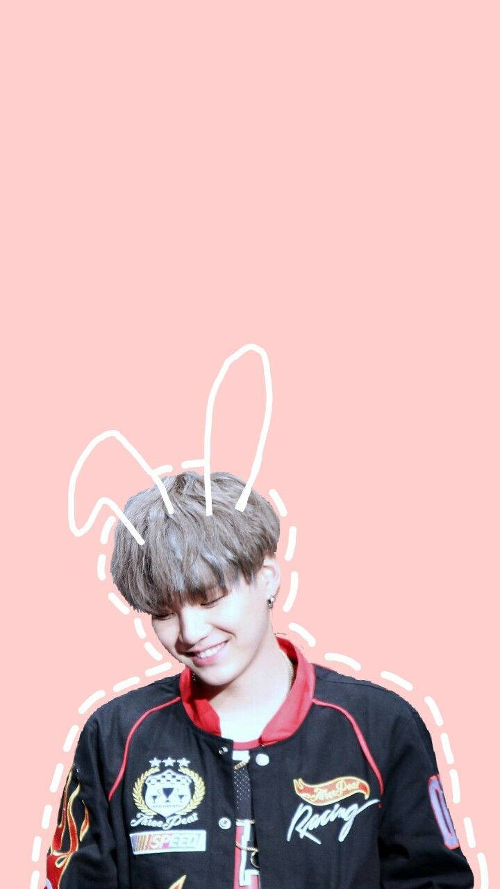 BTS 鎖定螢幕 桌布 壁紙 — 一個年輕男子帶有兔耳塗鴉，在柔和的粉紅色背景上溫暖地微笑。