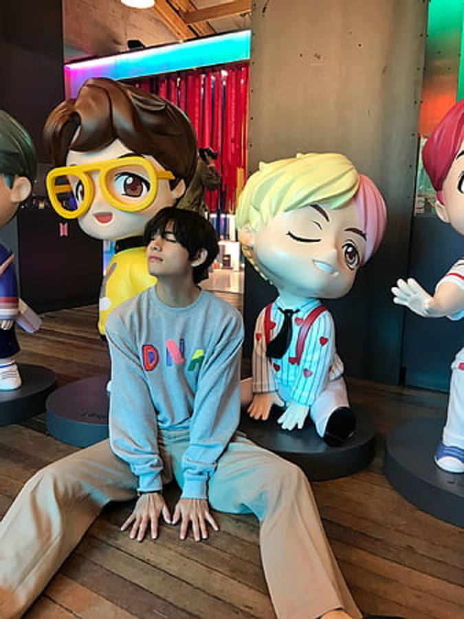 BTS 桌布 壁紙 — 一個年輕男子平靜地坐在木地板上，周圍是大型色彩繽紛的卡通雕像，處於充滿活力的室內環境中。