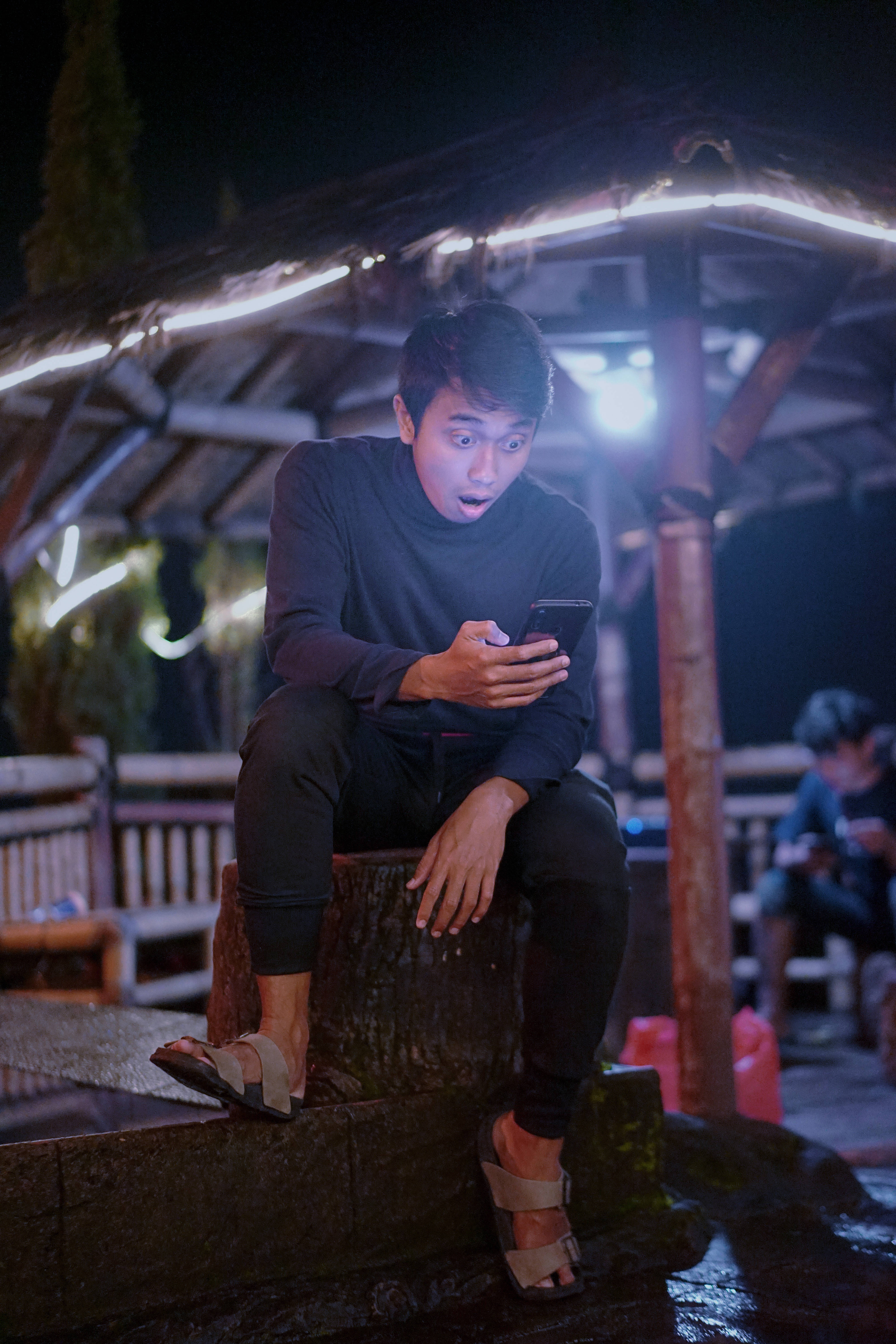 Cool Sad 桌布 壁紙 — 一個年輕男子晚上坐在發光的木製涼亭下的樹樁上，震驚地盯著手機。
