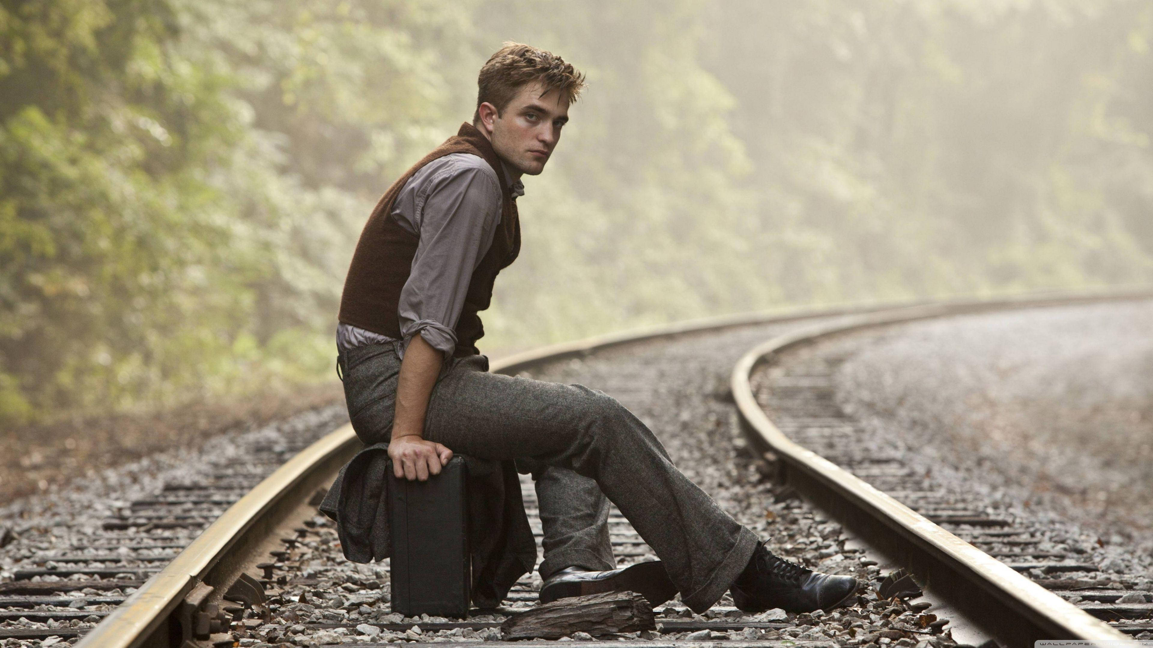 Robert Pattinson 桌布 壁紙 — 一個年輕男子沉思地坐在被霧氣籠罩的綠色森林中的火車軌道上。