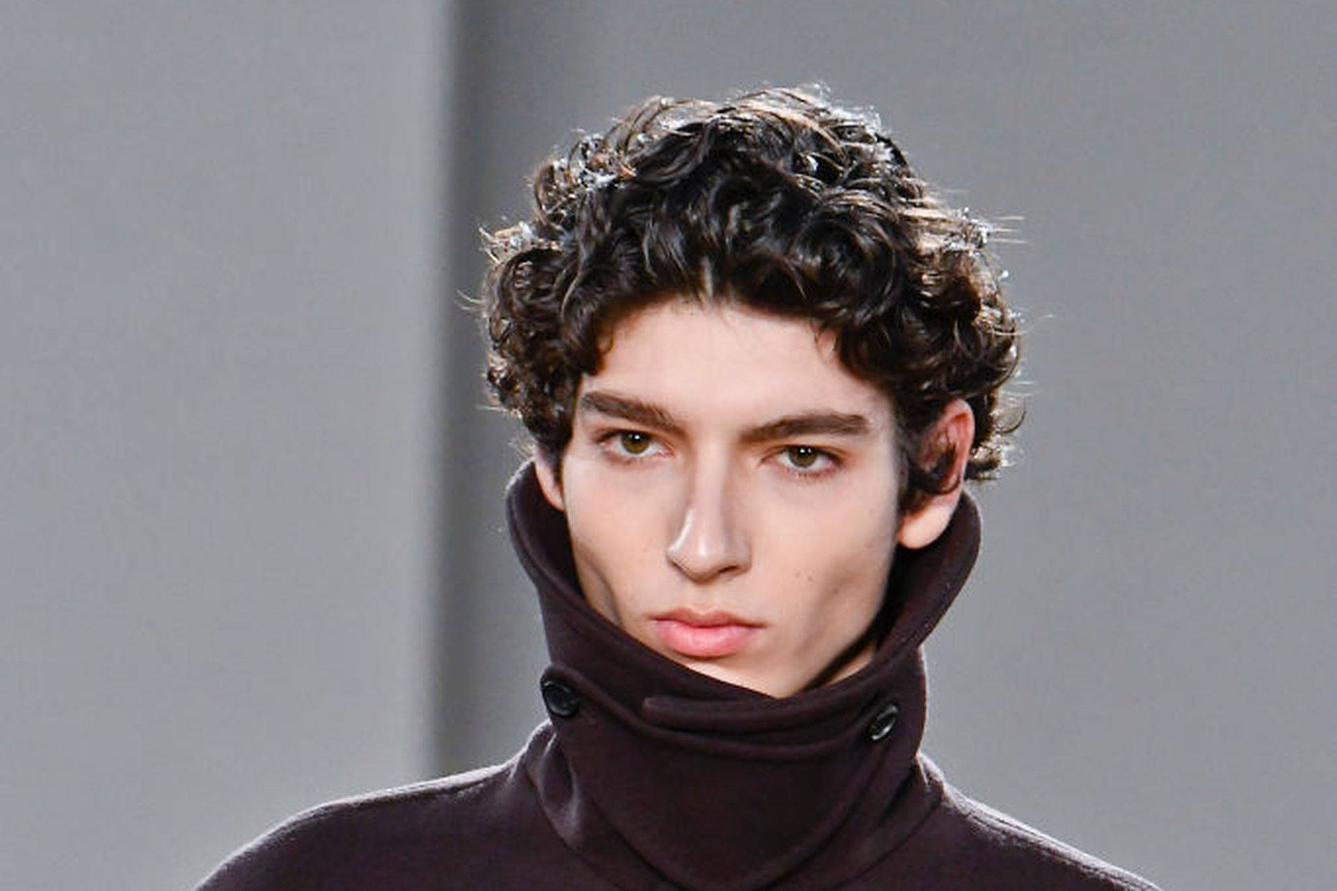 Ferragamo 桌布 壁紙 — 一個年輕男子的特寫，他有捲曲深色頭髮，穿著一件高領深棕色毛衣，背景是純灰色。