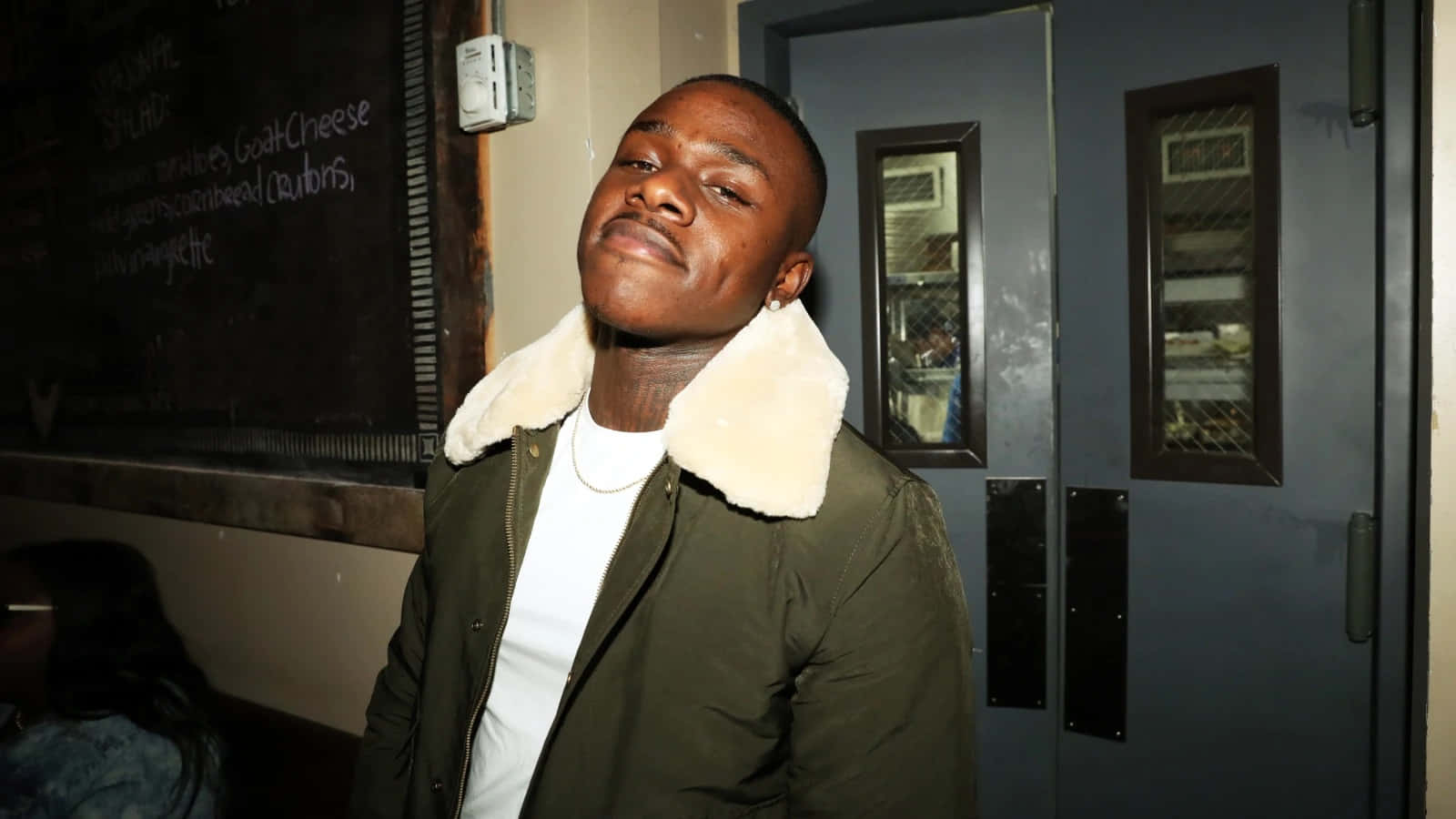 Dababy 桌布 壁紙 — 一個年輕男子穿著綠色夾克，白色毛領，在夜晚昏暗的門口自信地站立。