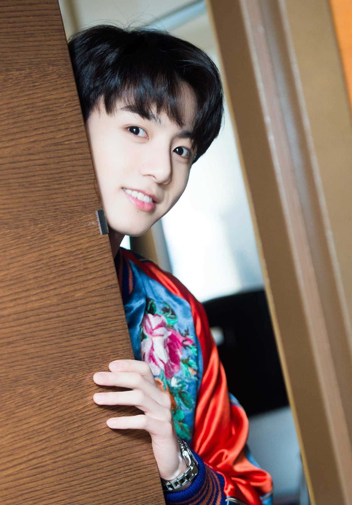 Jungkook 桌布 壁紙 — 一個年輕男子穿著鮮豔的花卉緞面夾克，俏皮地從木門後面探出頭來。