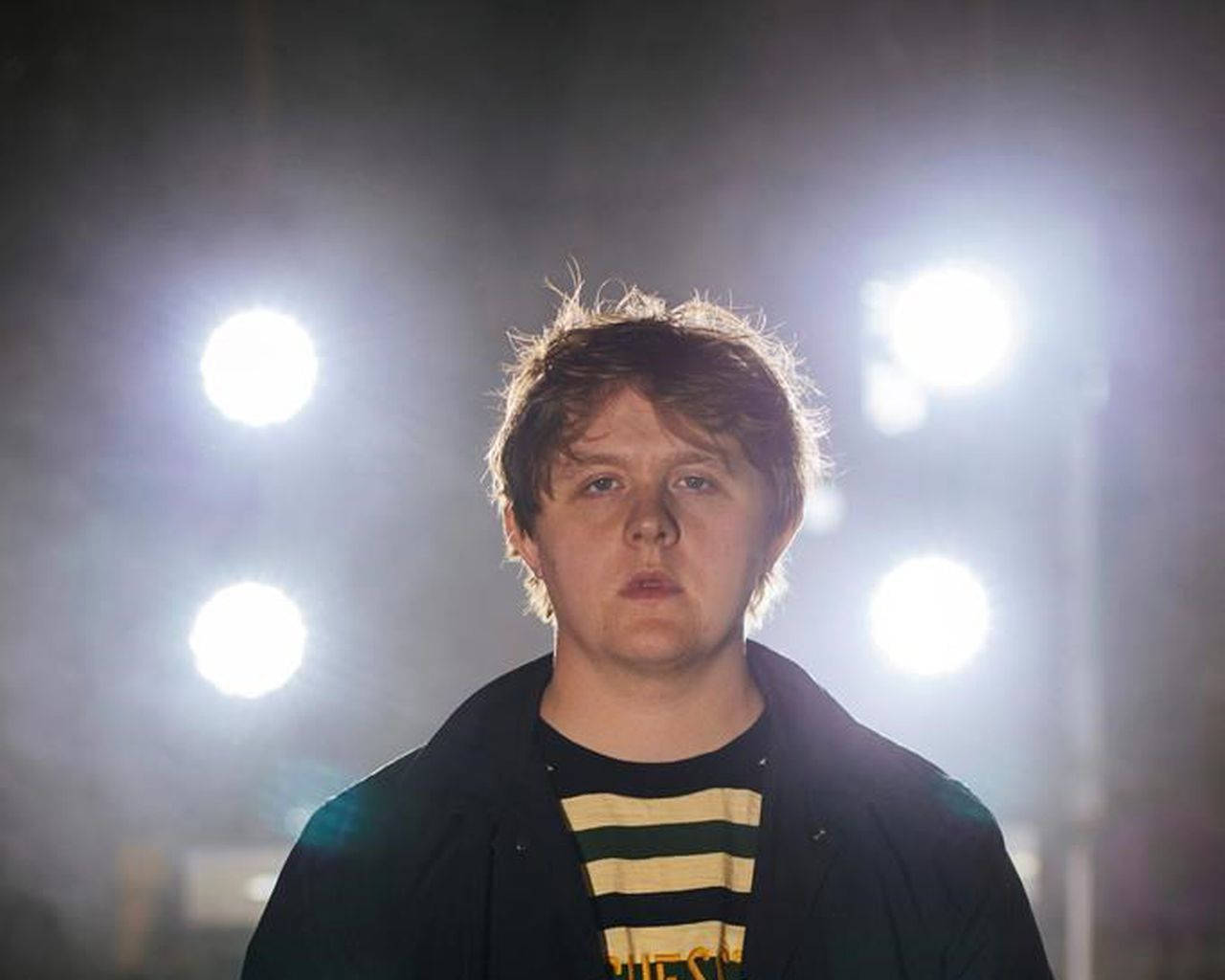 Lewis Capaldi 桌布 壁紙 — 一個年輕男子站在明亮的舞台燈光下，表情嚴肅，身處迷濛的氛圍中。