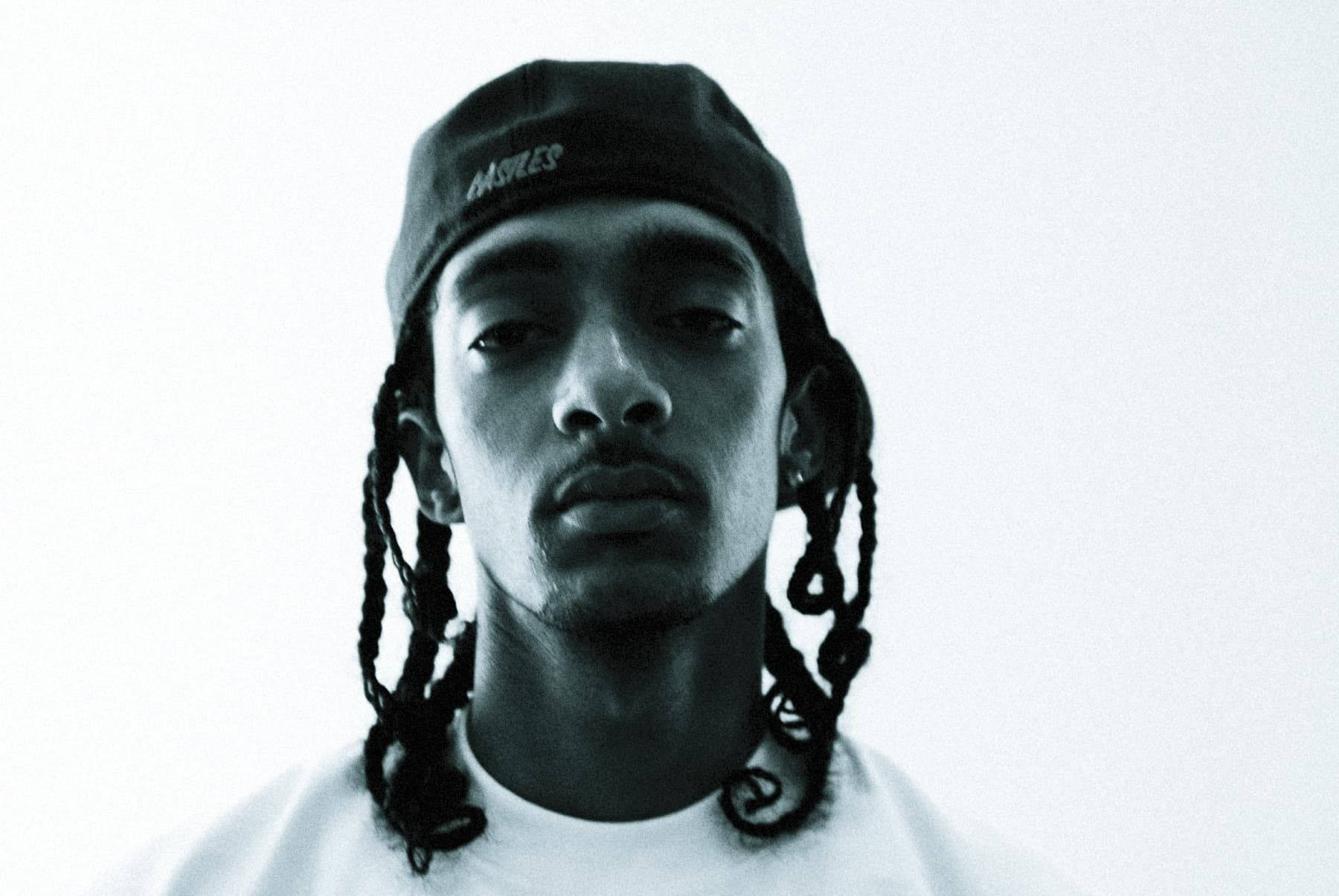 Nipsey Hussle 桌布 壁紙 — 一個年輕男子編織頭髮戴著帽子，在純色背景下的單色肖像。