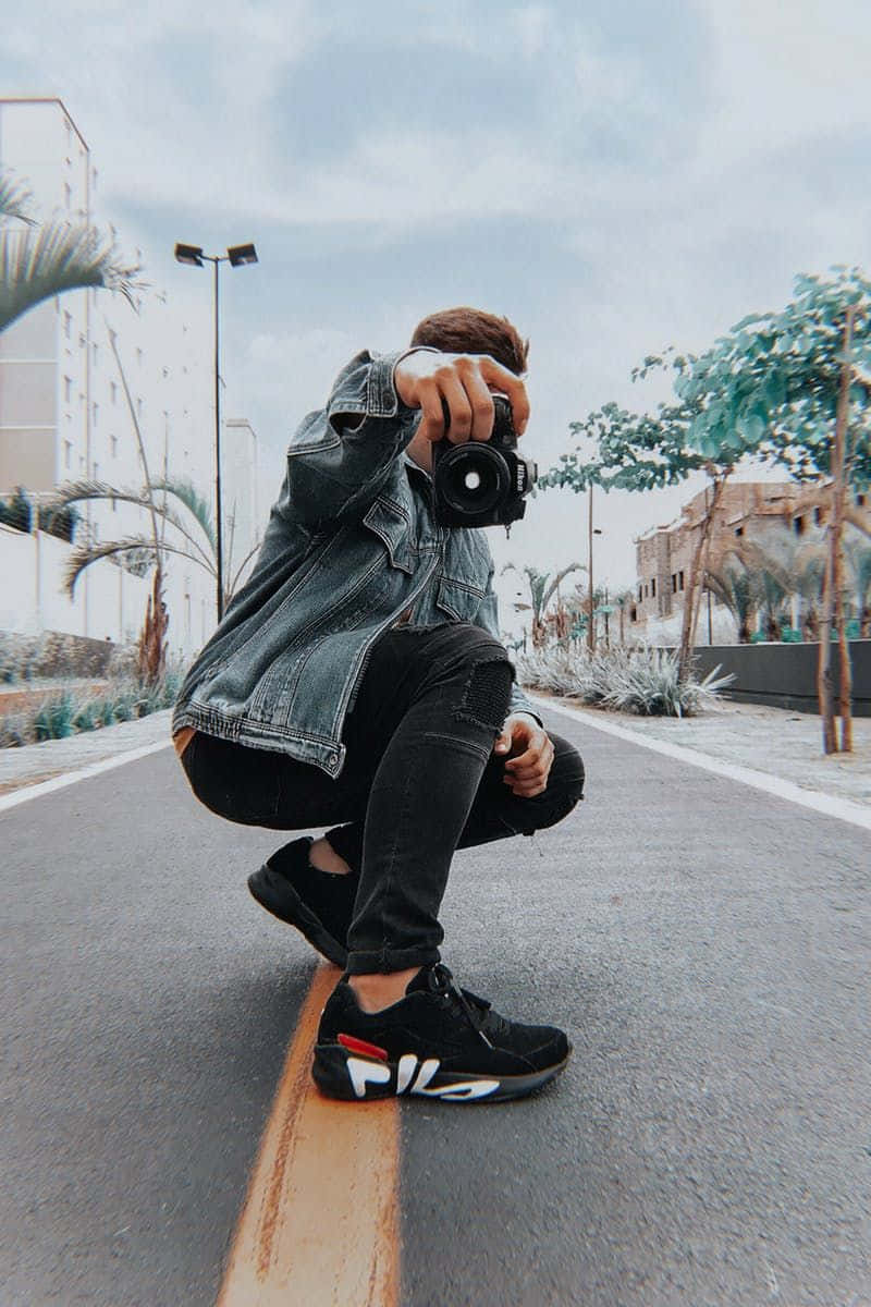 Holding Camera 桌布 壁紙 — 一個年輕男子蹲在鋪砌的道路上，手持相機，身穿牛仔外套和黑色運動鞋。