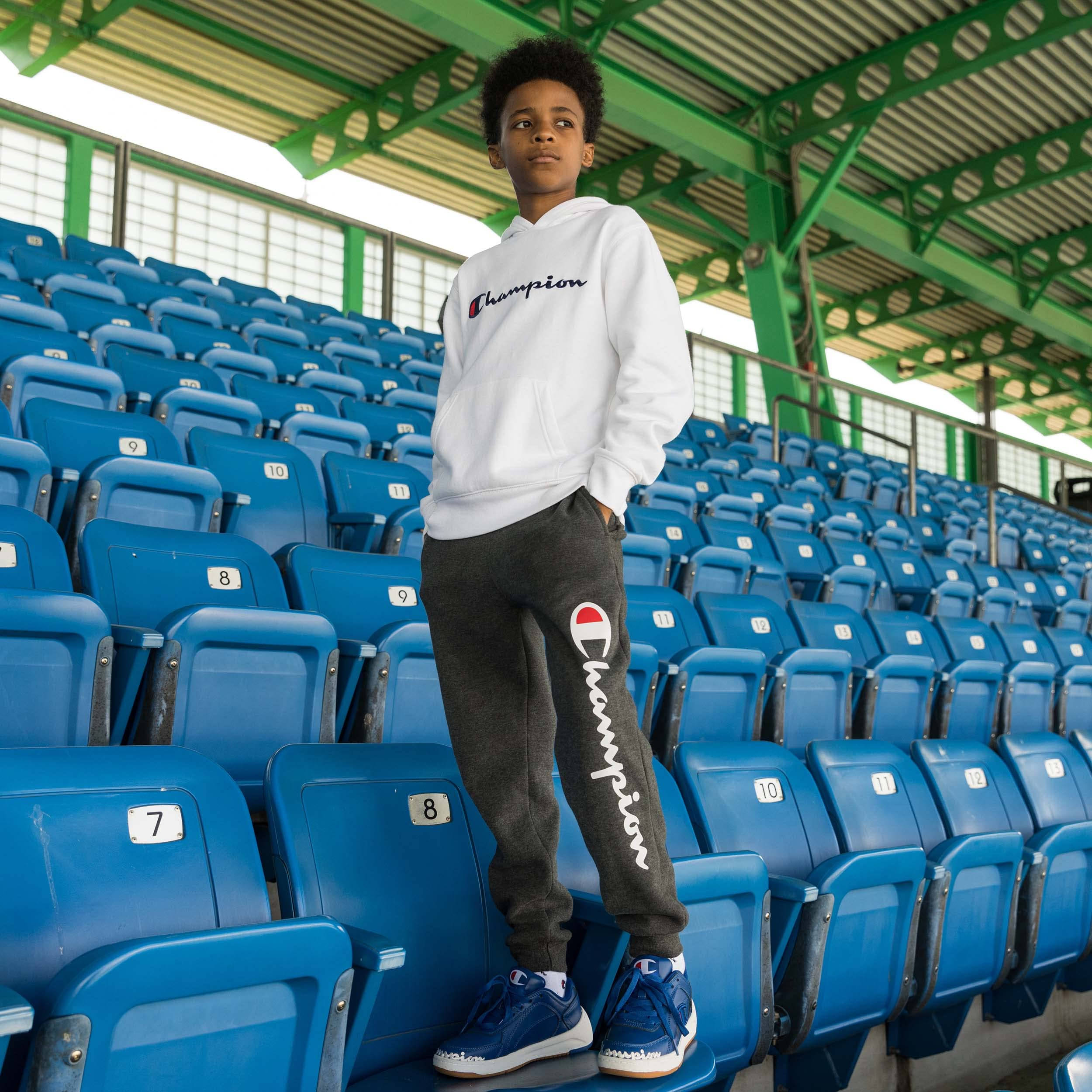 Champion 桌布 壁紙 — 一個年輕男孩穿著Champion連帽衫和運動褲，自信地站在藍色體育場座位上，上方是綠色金屬屋頂。