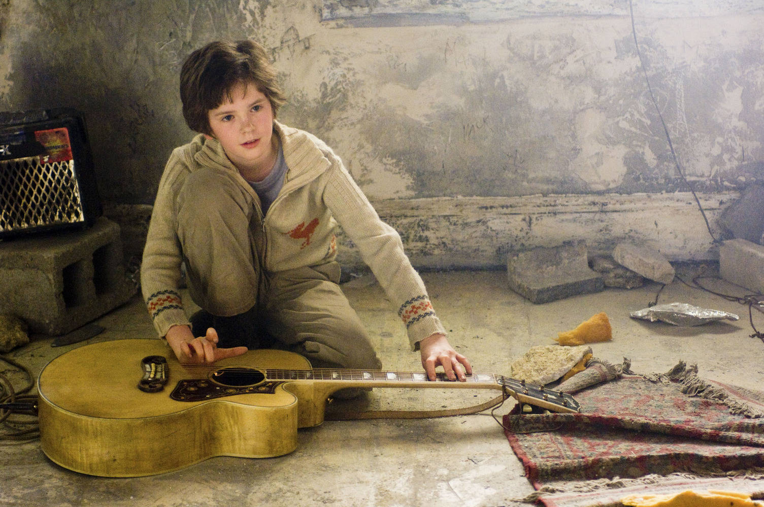 Freddie Highmore 桌布 壁紙 — 一個年輕男孩跪在一把破舊的原聲吉他旁邊，身處一個佈滿灰塵、被遺棄的房間，牆壁剝落，碎片散落一地。