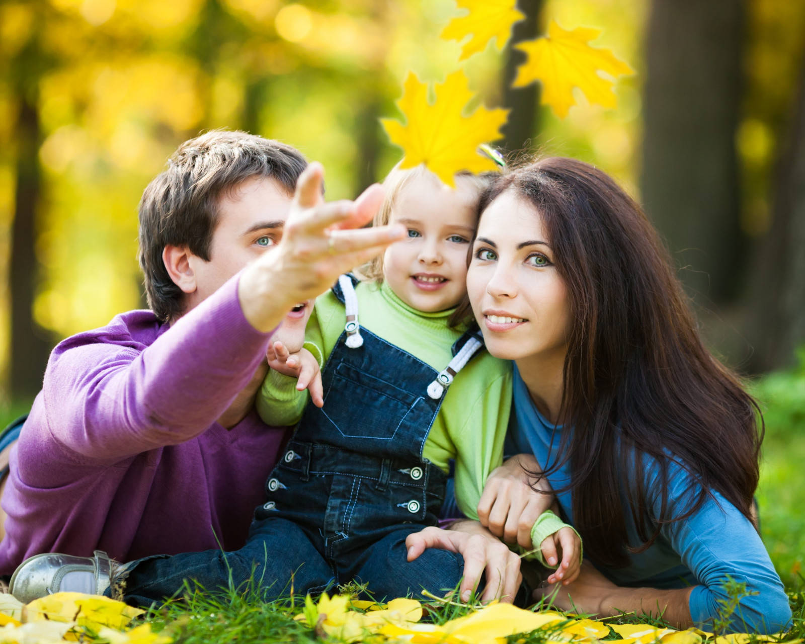 Mom 桌布 壁紙 — 一個幸福的三口之家在陽光明媚的秋日森林中一起大笑，金色的樹葉在他們周圍飄舞。