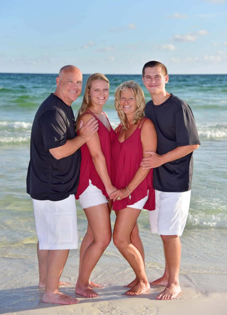 Beach Family 桌布 壁紙 — 一個幸福的四人家庭赤腳站在陽光明媚的海灘上，身後是溫柔的海浪和清澈的藍天。